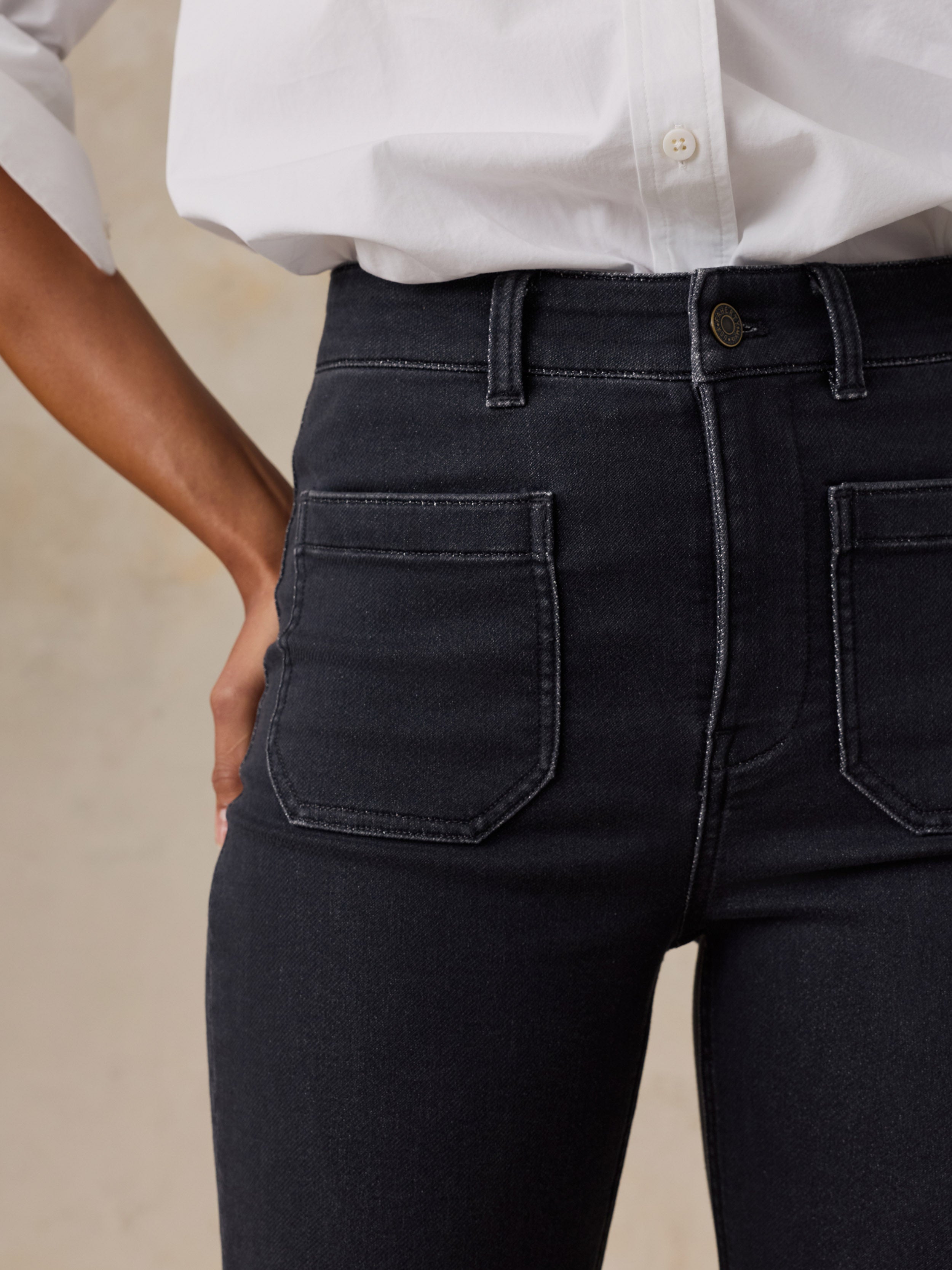 Close-up of a person wearing dark charcoal black high-waisted jeans with a white button-down shirt, shown from the waist down against a warm beige background.