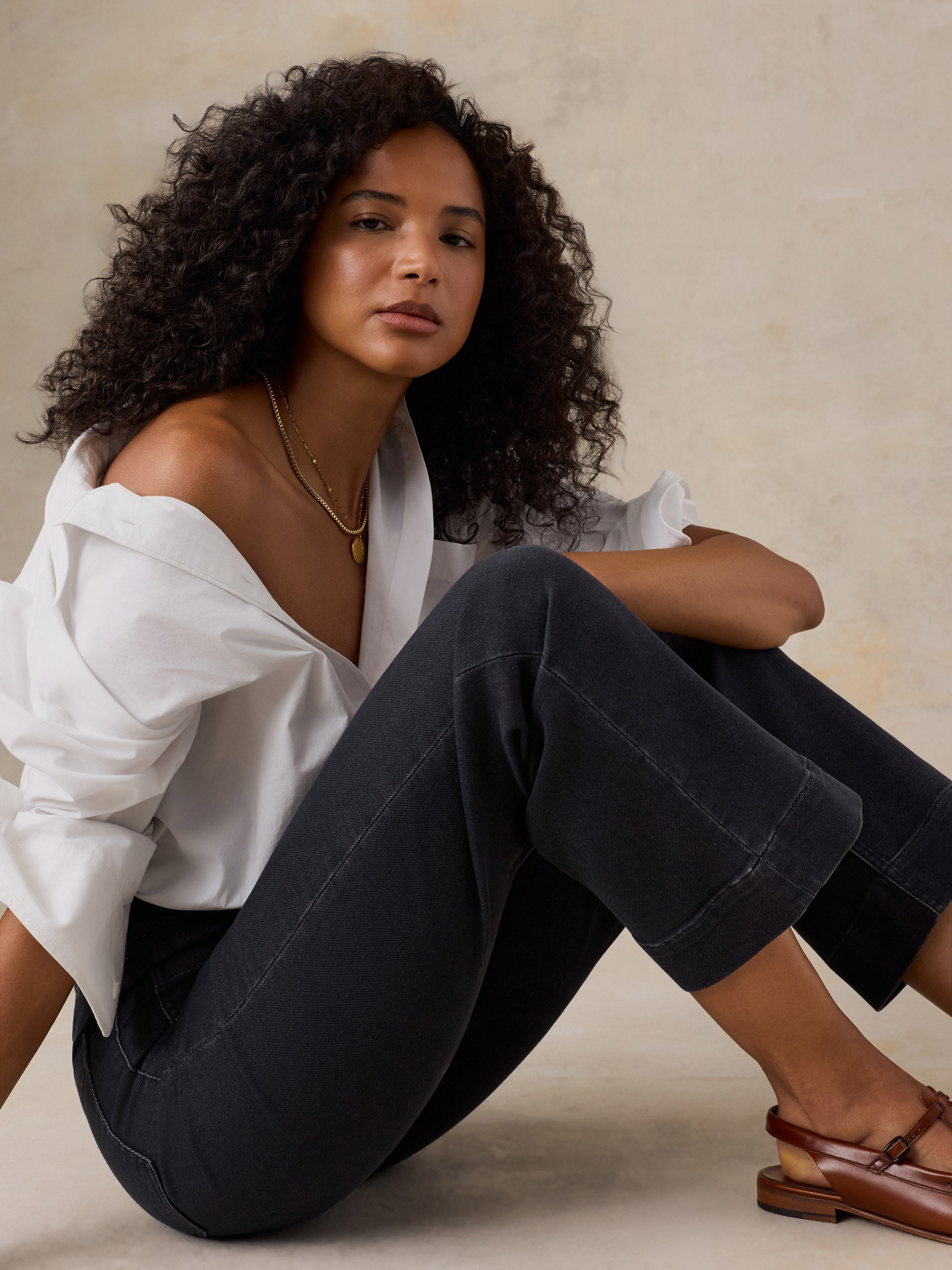 A woman with curly dark hair sits cross-legged on a beige floor against a textured beige wall, wearing a white button-down shirt, dark charcoal cropped pants, and brown leather loafers.