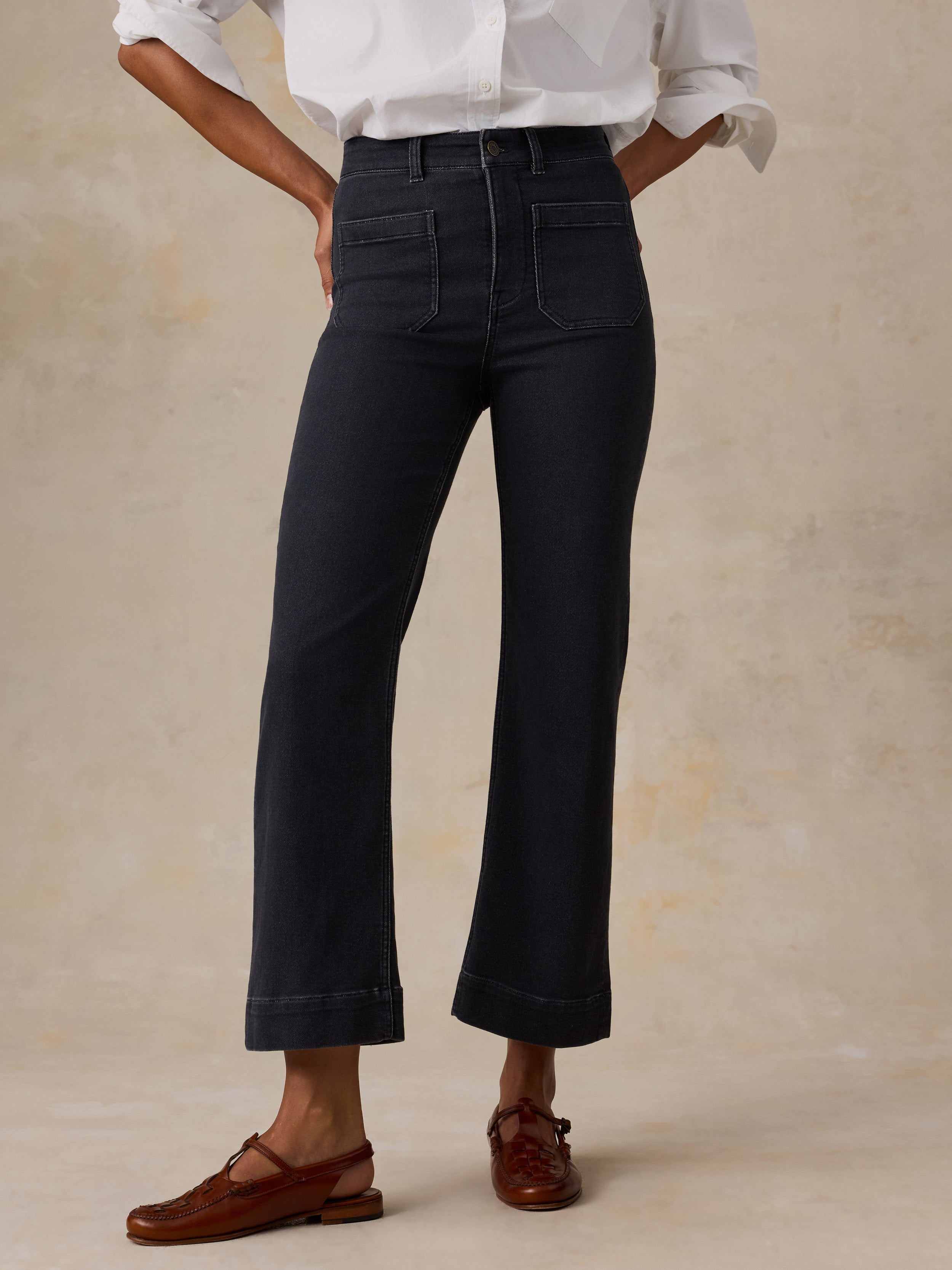 A model wearing black high-waisted cropped flare jeans, a white button-down shirt, and brown leather loafers, standing against a textured beige studio backdrop.