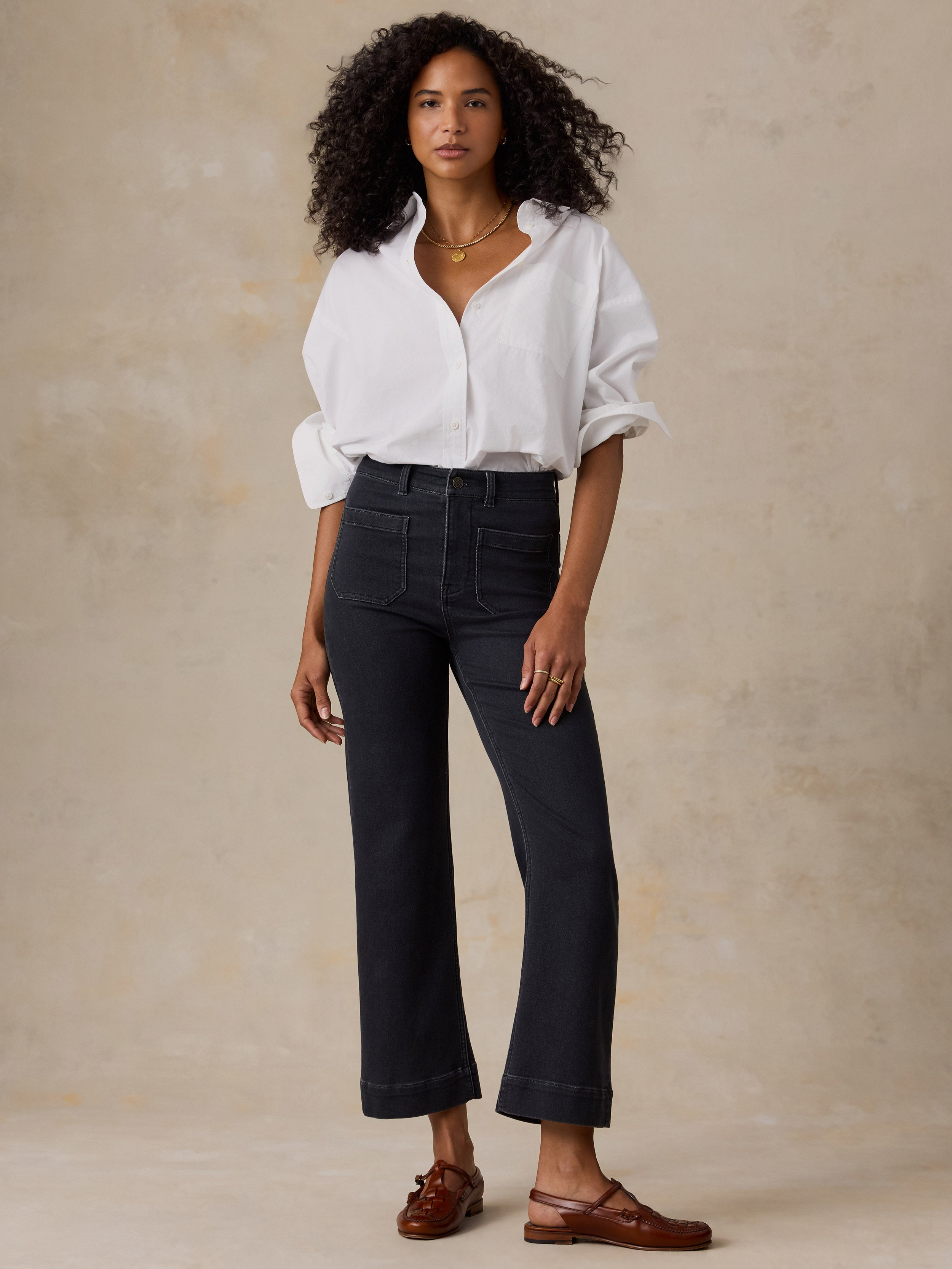 A woman modeling a white button-down shirt and black wide-leg cropped jeans, paired with black loafers, against a warm beige studio backdrop.