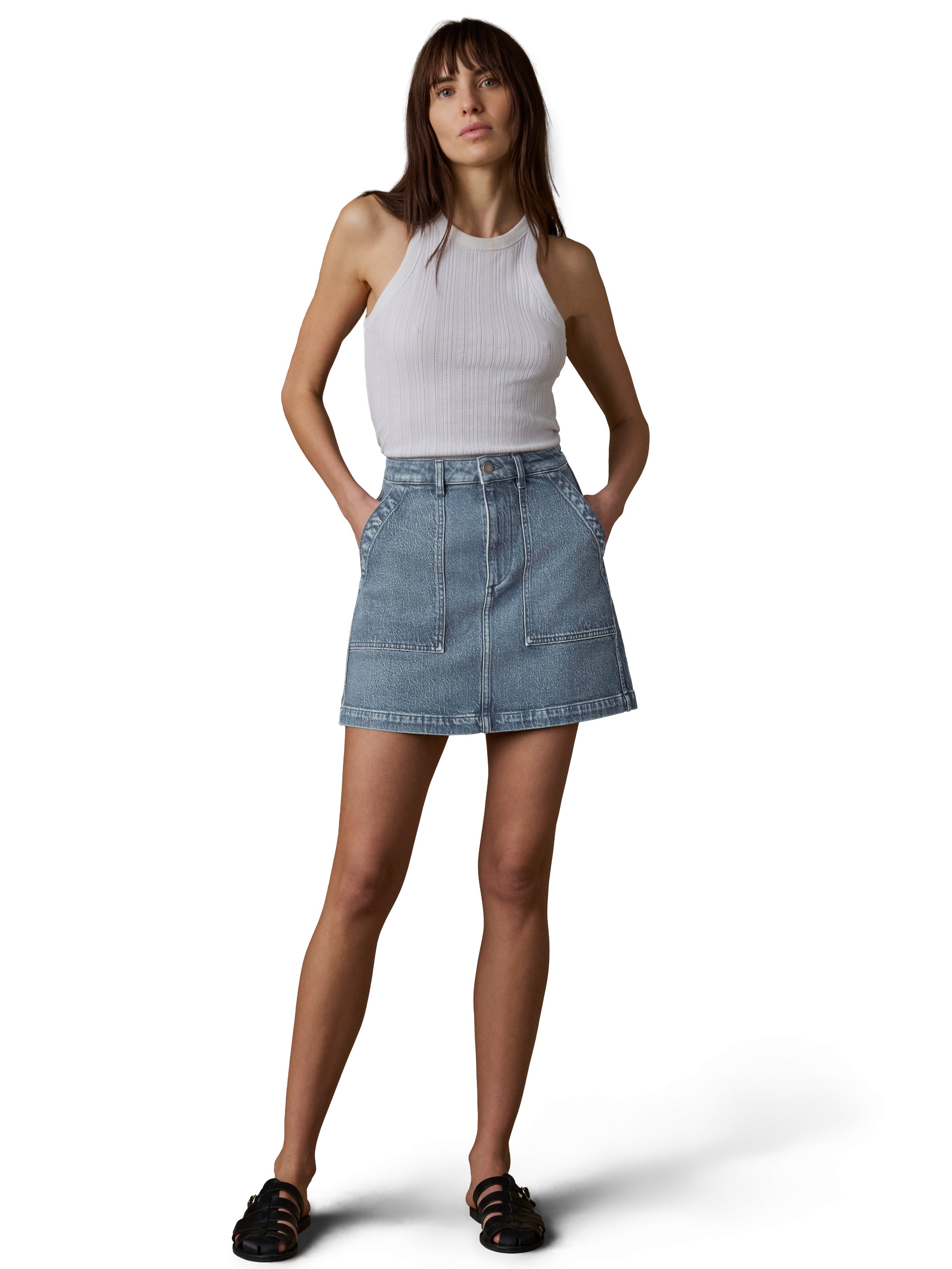 A woman wearing a white tank top, denim skirt, and black sandals