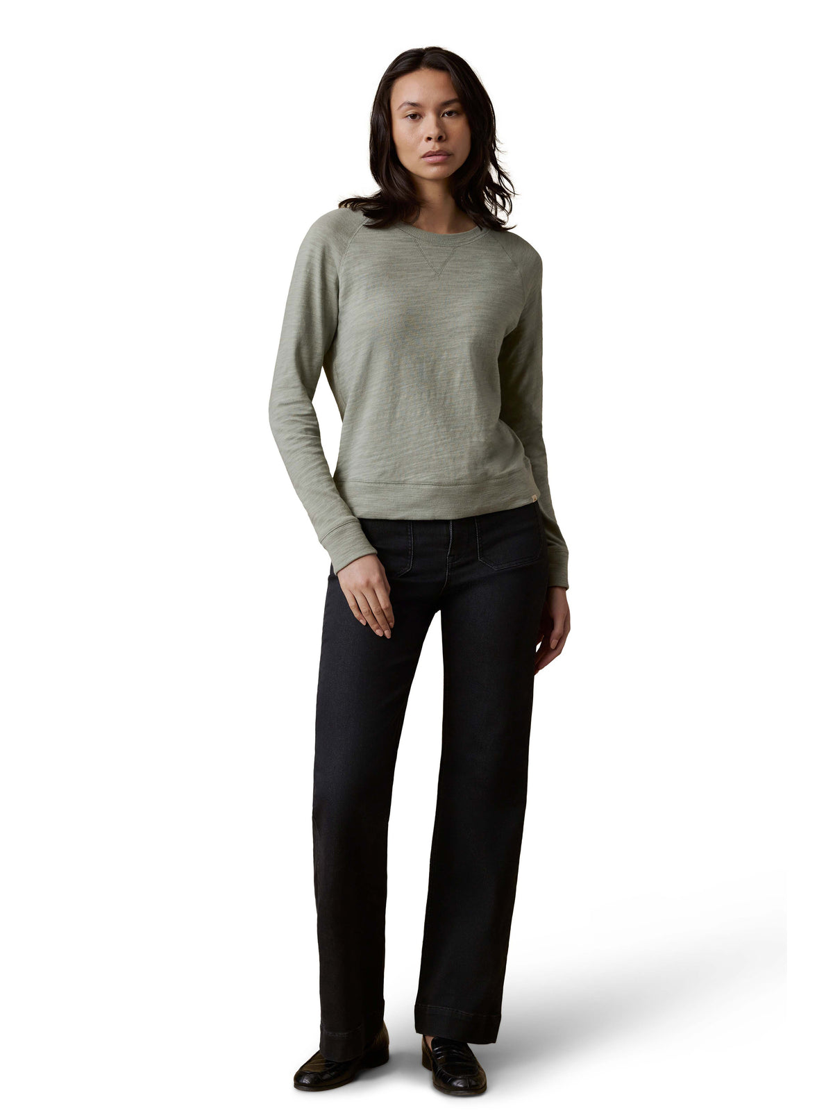 A woman wearing a sage green crewneck, dark jeans, and loafers, image 2 of 3