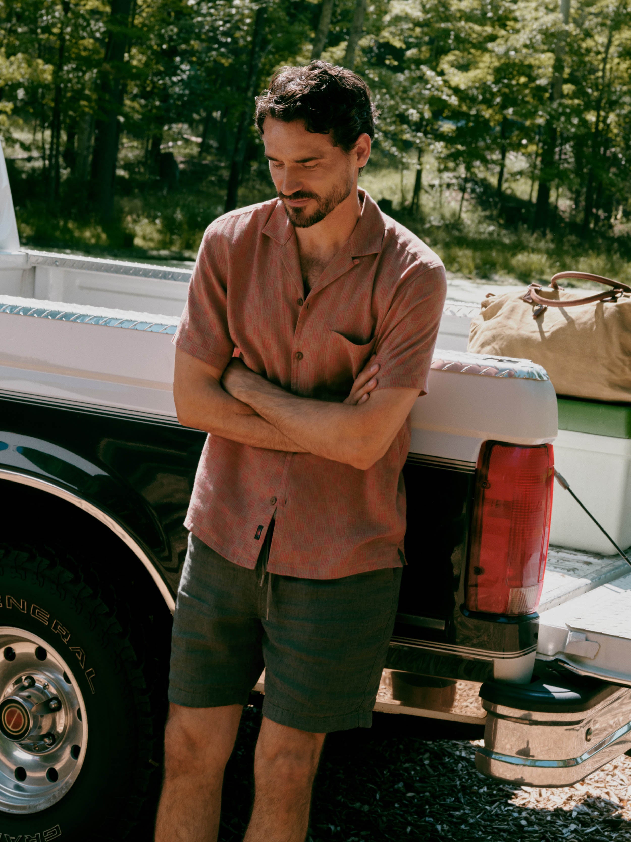 A man stands leaning against the back of a white pickup truck outdoors, wearing a muted dusty rose short-sleeve button-up shirt and dark olive-gray shorts. Trees and gravel are visible in the background, along with a tan duffel bag in the truck bed.