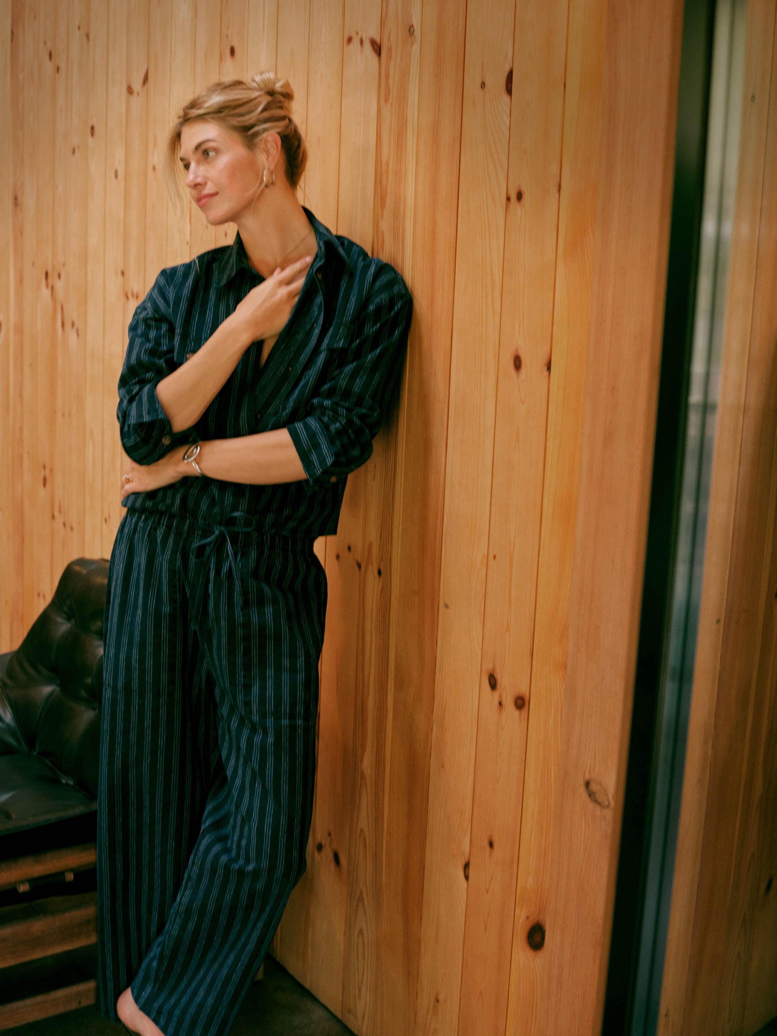 A woman wearing a dark navy blue and light blue vertically striped shirt and matching pants leans against a warm wood-paneled wall.
