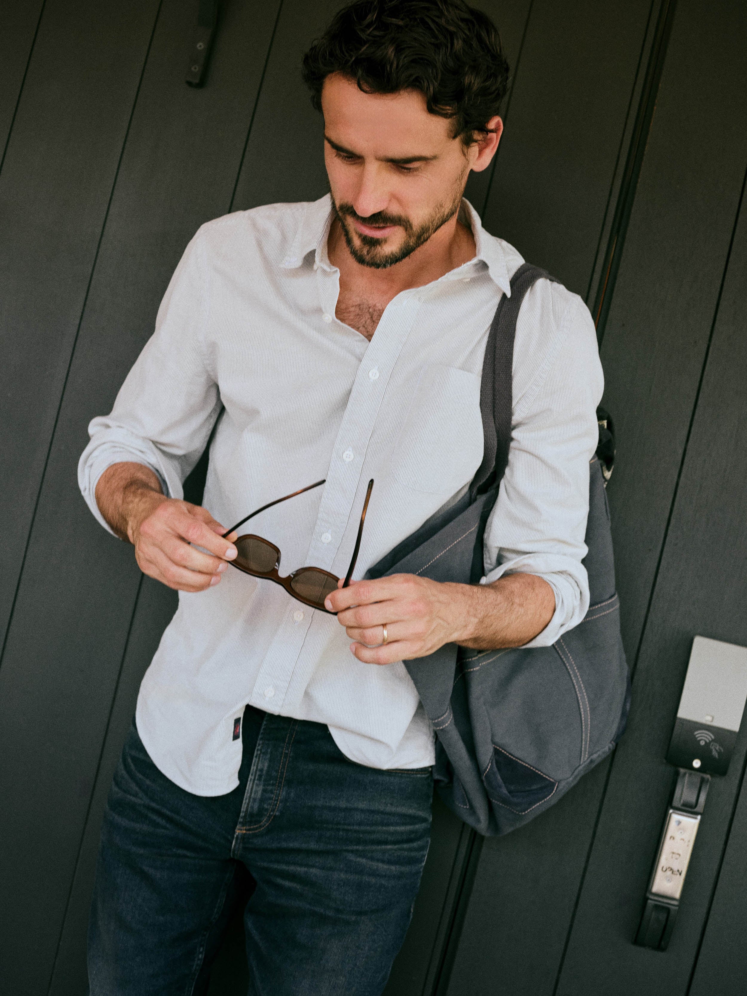 A man wearing a white and blue striped button-down shirt and dark blue jeans stands against a dark wood-paneled wall, holding a pair of sunglasses in his hands while carrying a gray shoulder bag.