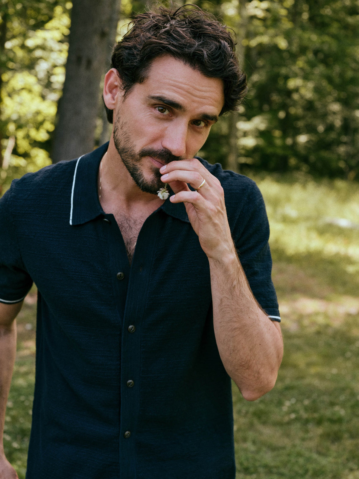 A man wearing a dark navy blue short-sleeve polo shirt with white piping on the collar and sleeve cuffs, standing outdoors in a grassy, wooded area., image 1 of 6