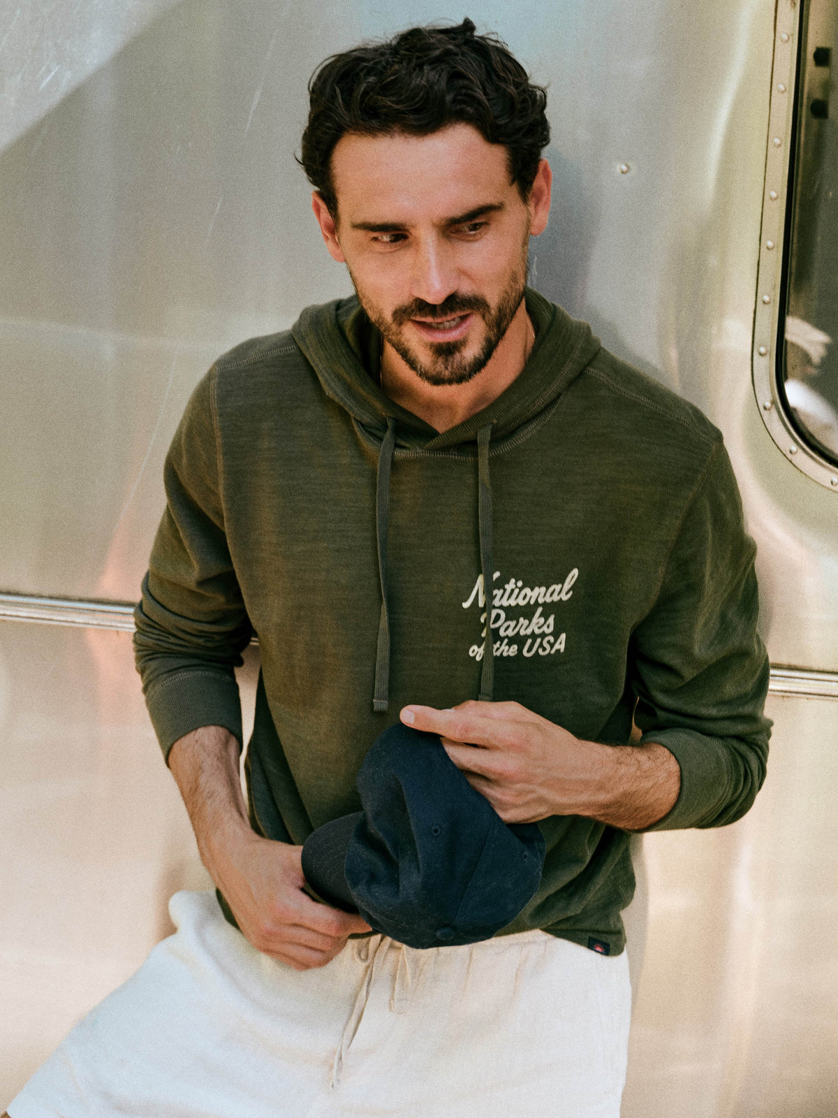 A man wearing an olive green hoodie with 'National Parks of the USA' printed in white script on the chest, paired with white sweatpants and holding a dark blue cap, posed in front of a metallic silver trailer., image 1 of 8