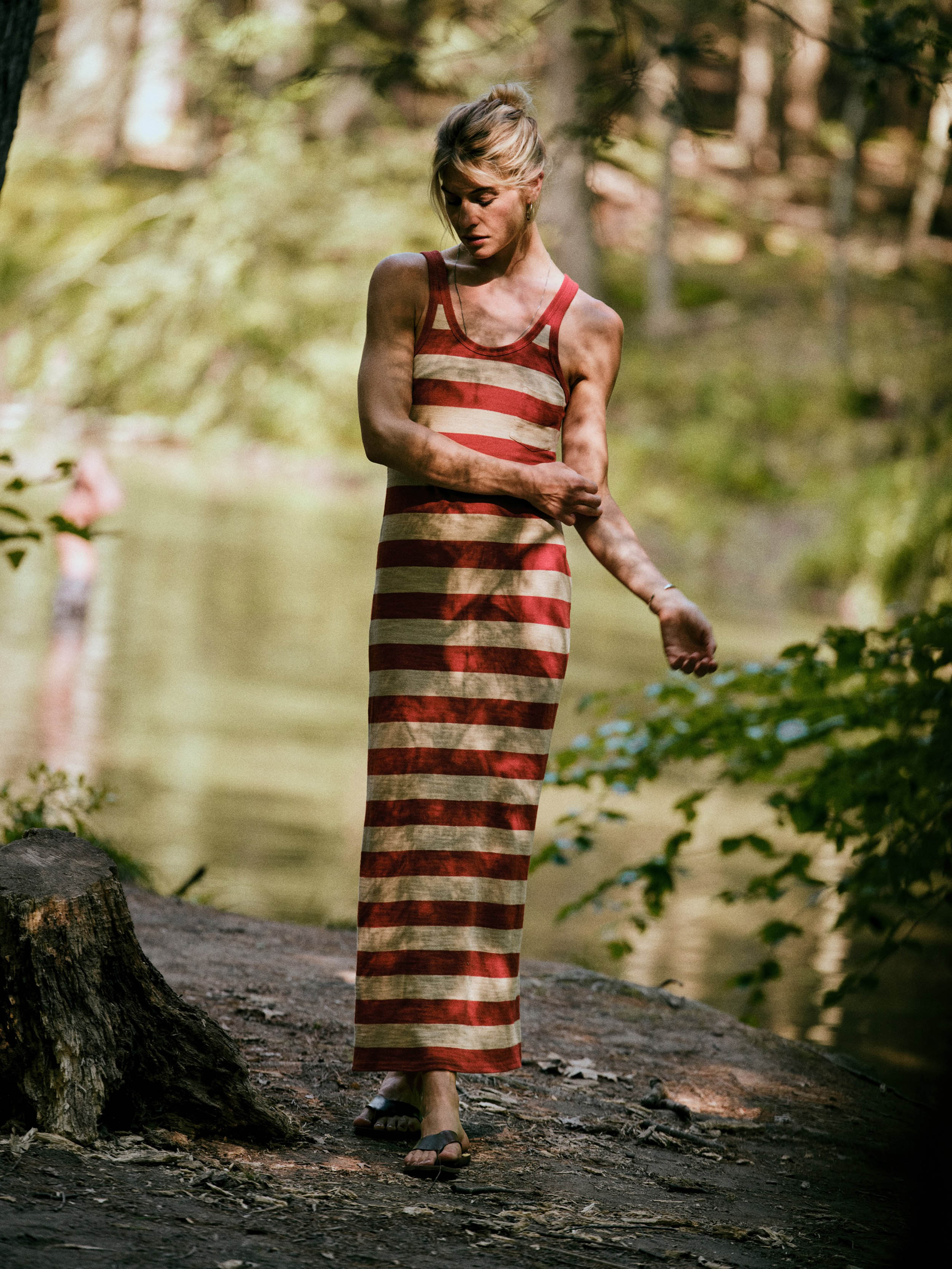 A woman wearing a red and beige horizontally striped maxi dress stands on a dirt path near a body of water in a wooded outdoor setting.