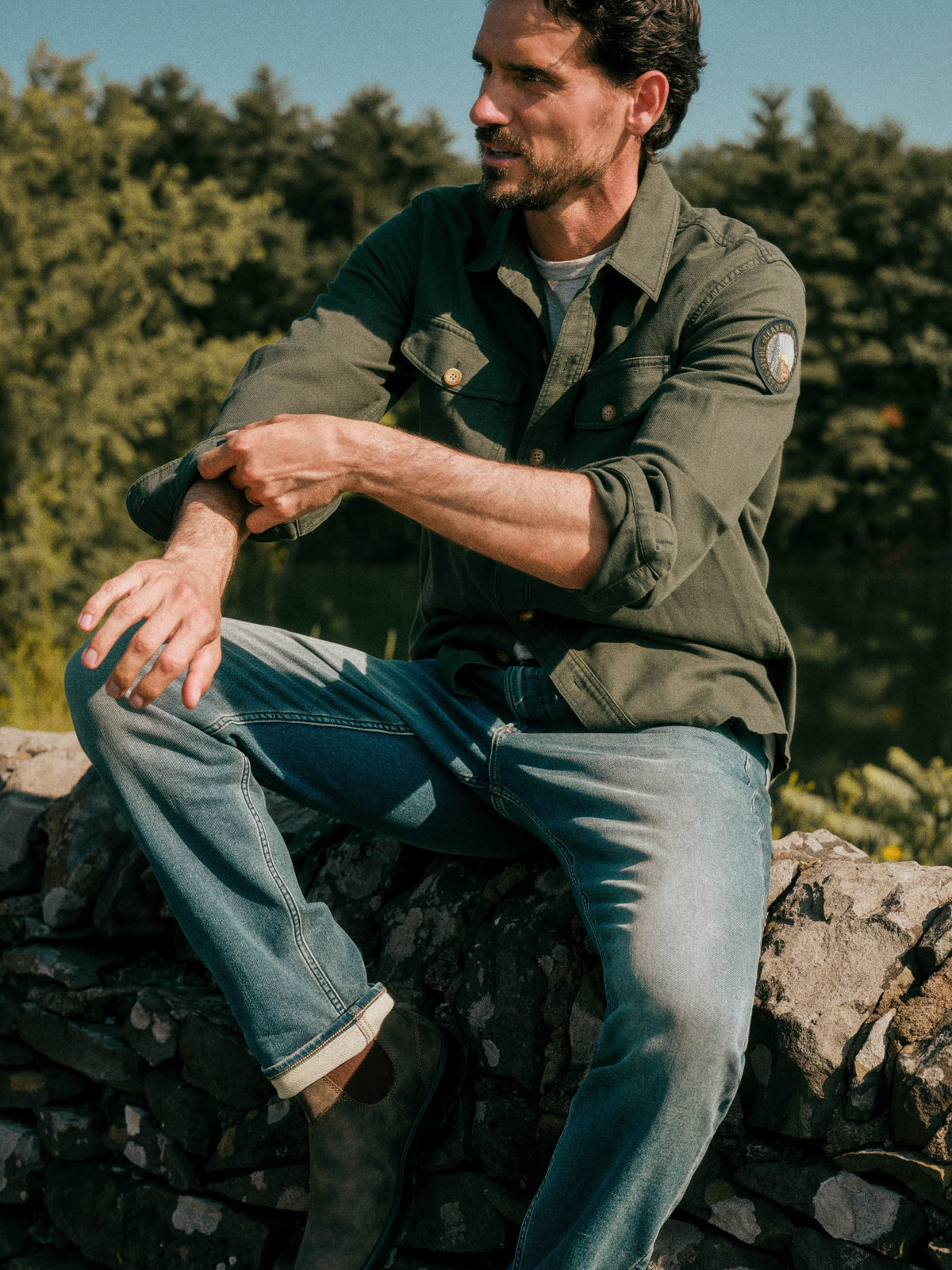 A man sitting outdoors on a stone wall wearing an olive green button-up shirt and blue jeans, with trees and blue sky in the background., image 1 of 10