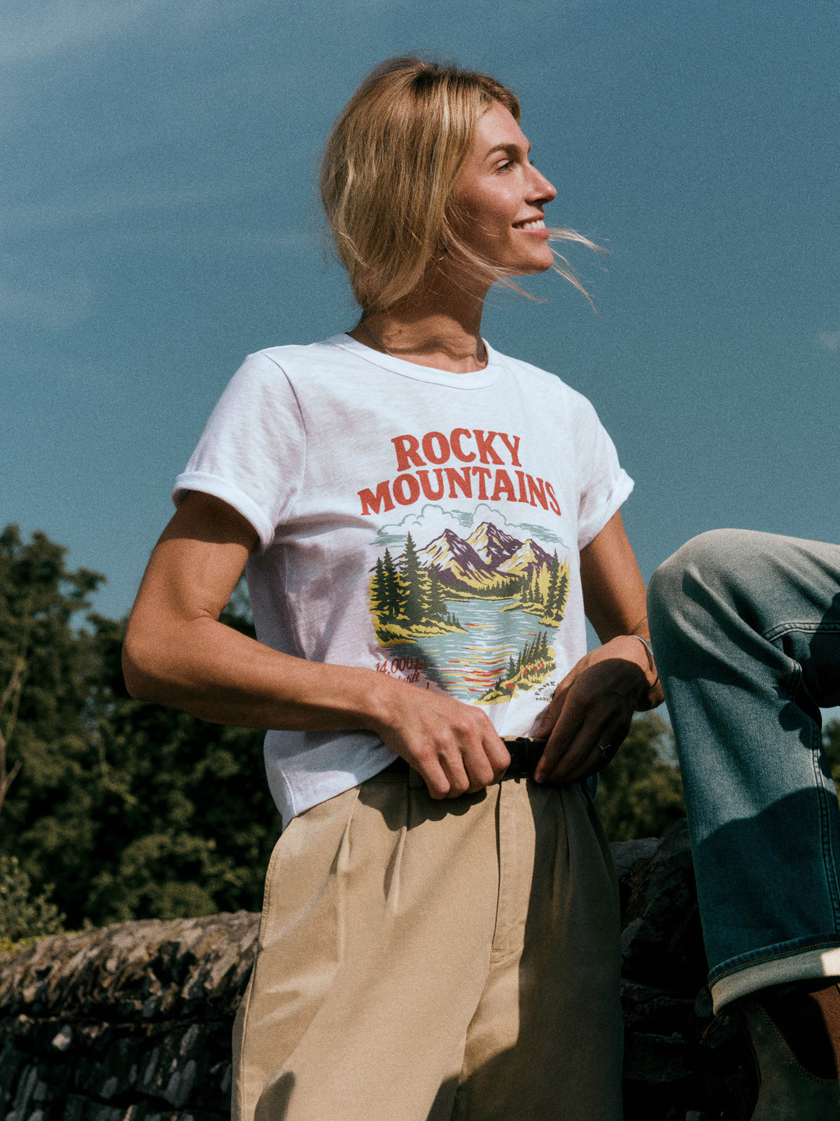 A woman wearing a white graphic t-shirt with 'ROCKY MOUNTAINS' and a mountain lake print, paired with beige pants, photographed outdoors against a blue sky., image 1 of 7