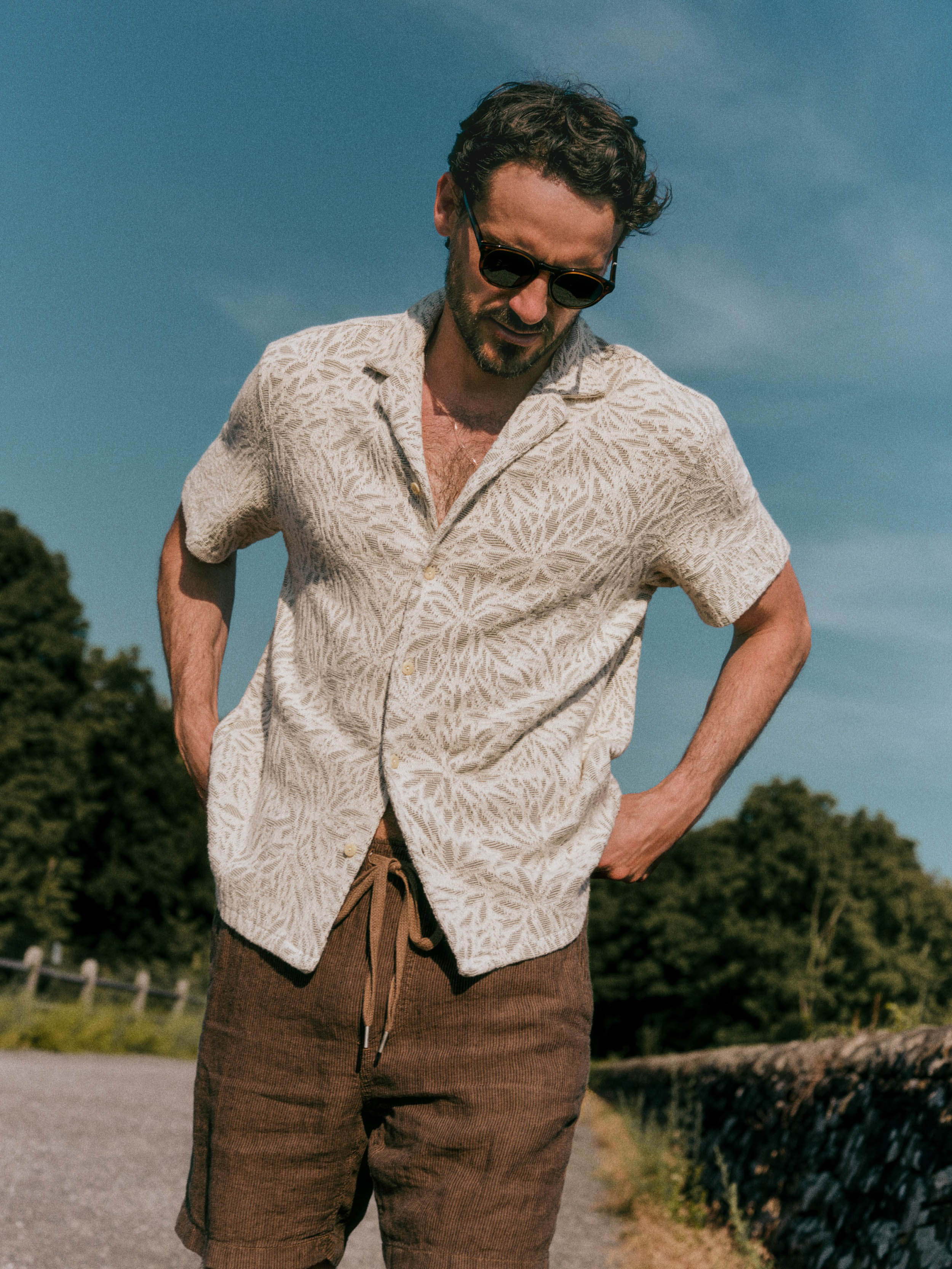 A man wearing a short-sleeve button-up shirt with a light green and white leaf pattern, paired with brown pants, standing outdoors on a sunny day with trees and a blue sky in the background.