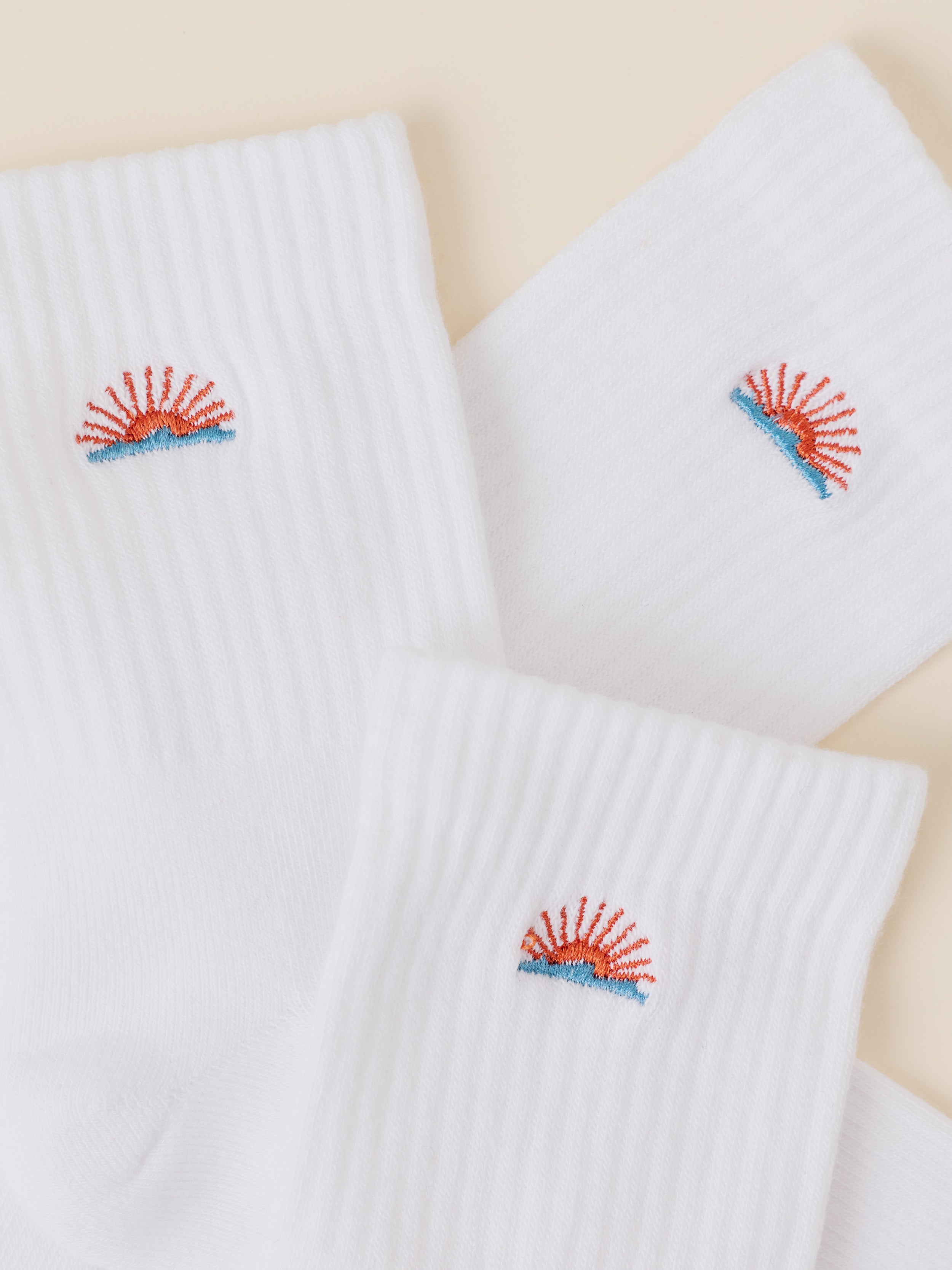Three white ribbed crew socks arranged on a light beige background, each featuring a small embroidered sun and waves graphic logo in red and blue near the cuff.