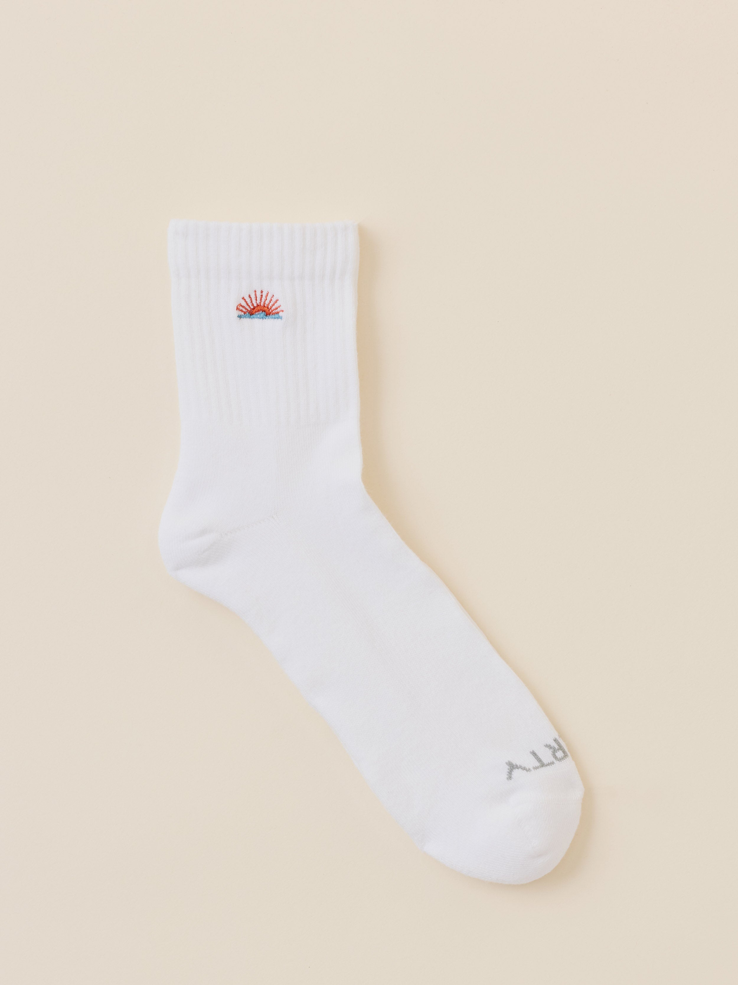 A single white crew sock with a small embroidered red and blue sun and waves graphic logo near the cuff and gray text near the toe, displayed on a solid beige background.
