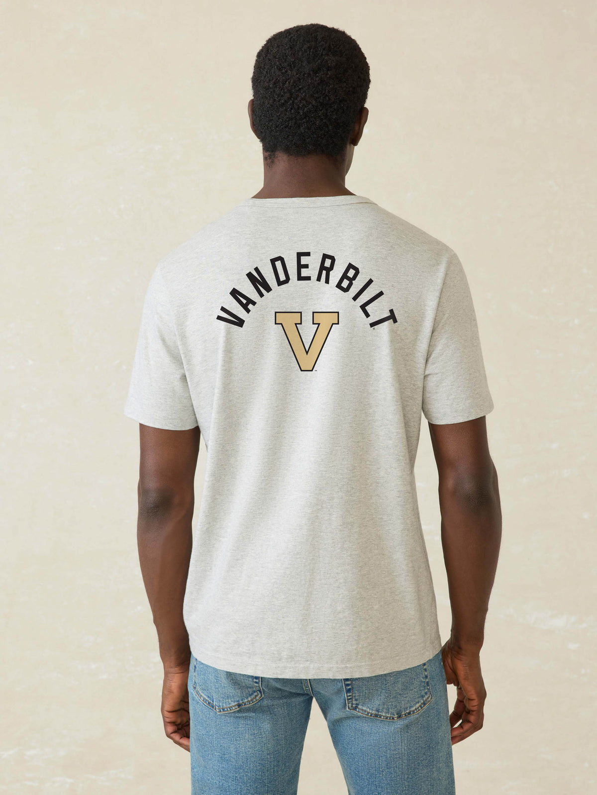 A man viewed from behind wearing a light heather gray t-shirt with a black and gold Vanderbilt logo on the back, paired with blue jeans, against a textured beige background., image 2 of 2