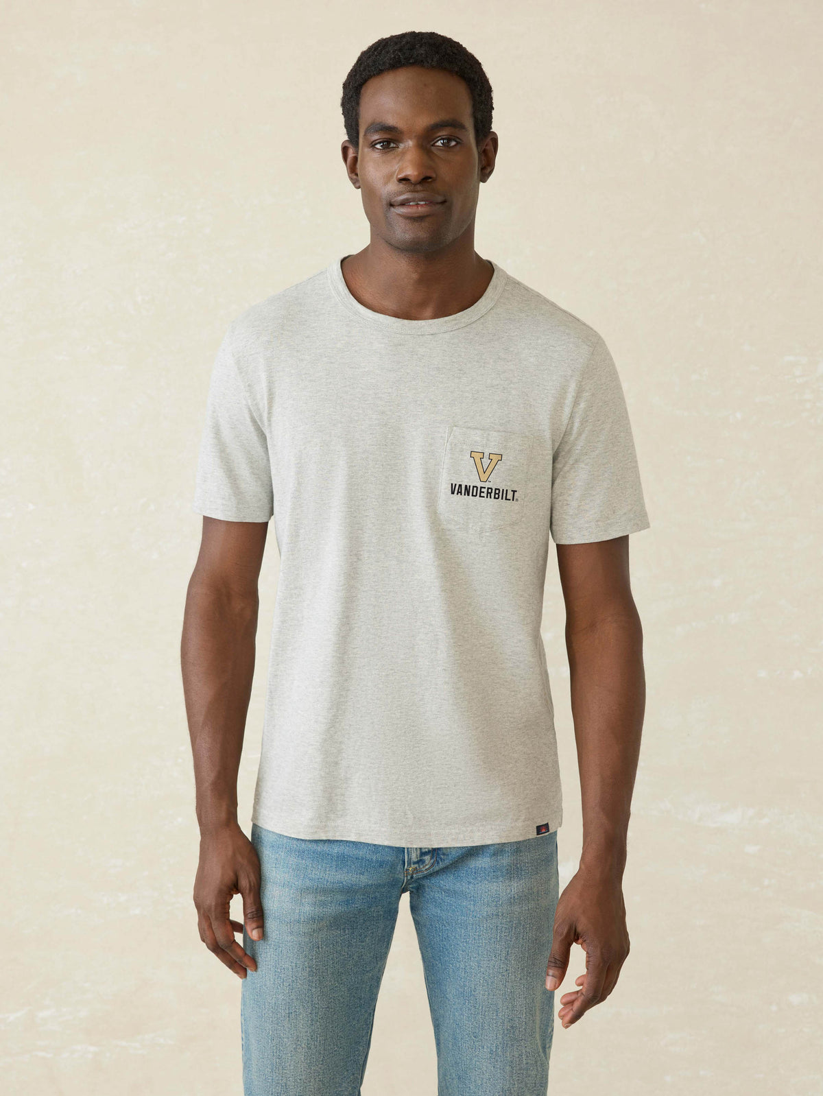 A man wearing a light heather gray t-shirt with a small gold and black 'Vanderbilt' logo on the left chest, paired with blue jeans, standing against a textured beige background., image 1 of 2
