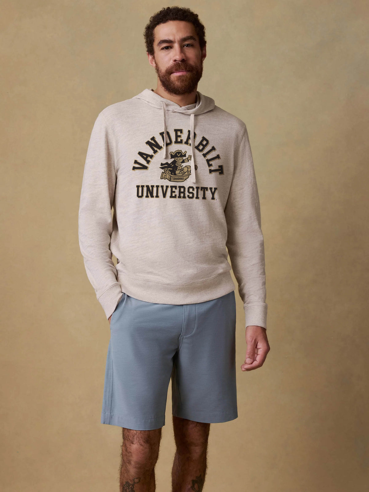 A man wearing a light beige heathered hoodie with a black 'VANDERBILT UNIVERSITY' graphic on the chest, paired with light blue shorts, standing against a textured beige studio backdrop., image 1 of 1