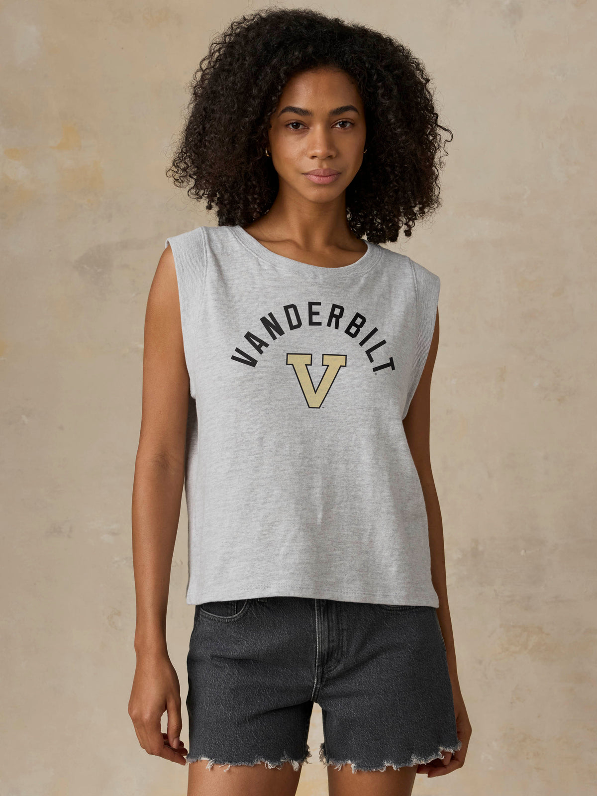 A woman modeling a light gray sleeveless crop top with 'VANDERBILT' and a large 'V' logo printed on the front, paired with dark denim shorts against a textured beige background., image 1 of 1