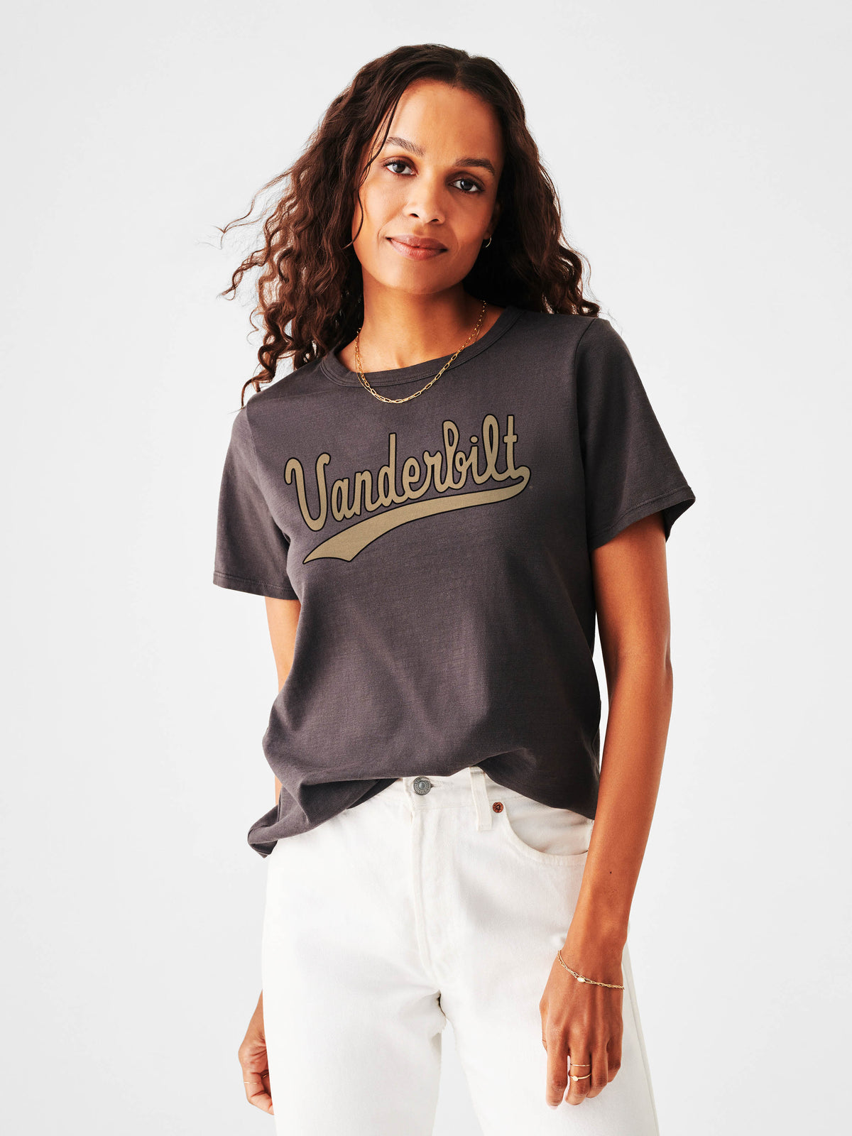 A woman wearing a dark gray t-shirt with a gold 'Vanderbilt' script graphic across the chest, paired with white pants, photographed against a light gray studio background., image 1 of 1