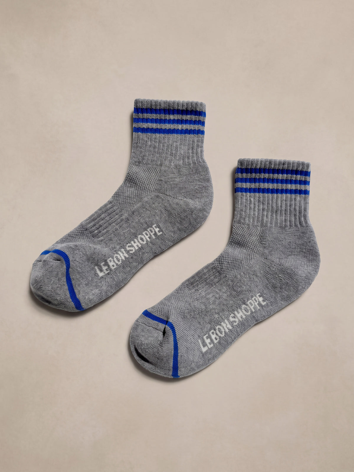 A pair of heather gray ankle socks with blue stripes on the cuff and toe, featuring the brand name 'Le Bon Shoppe' printed in white on the sole, displayed on a neutral beige background., image 2 of 2