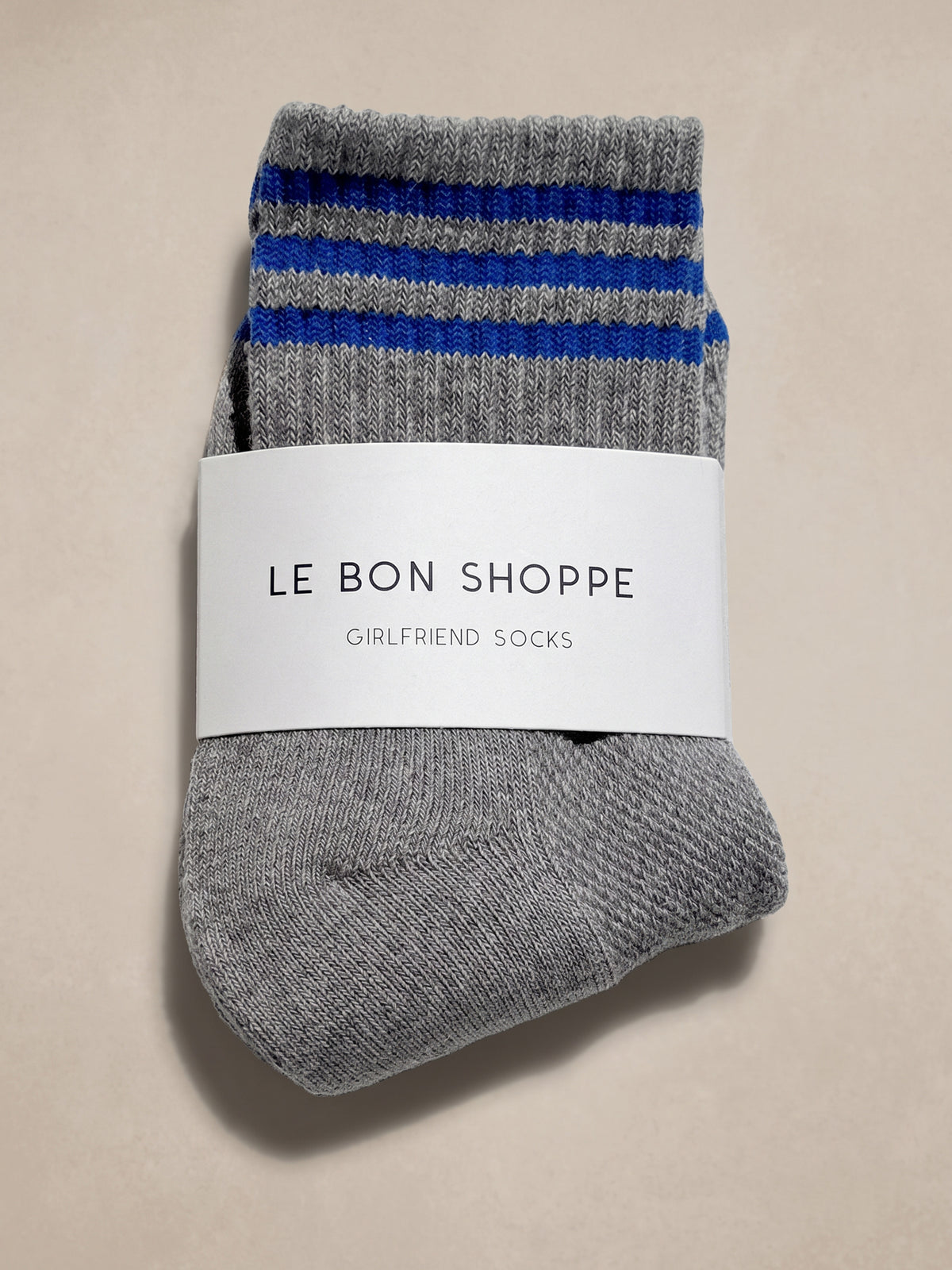 A pair of gray knit socks with blue and gray striped cuffs, wrapped in a white paper band that reads 'Le Bon Shoppe Girlfriend Socks', on a beige background., image 1 of 2