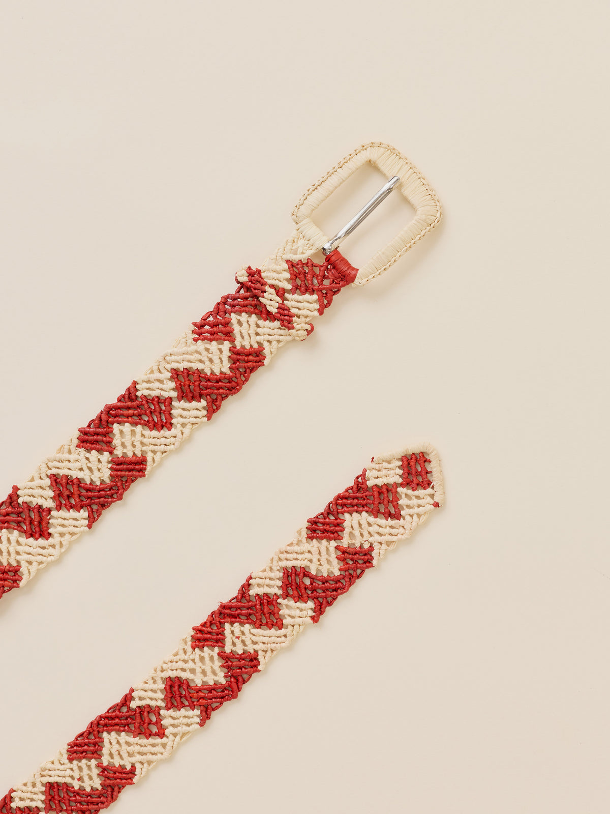 A woven braided belt with a red and cream chevron pattern and a silver metal buckle, shown on a solid light beige background., image 2 of 2