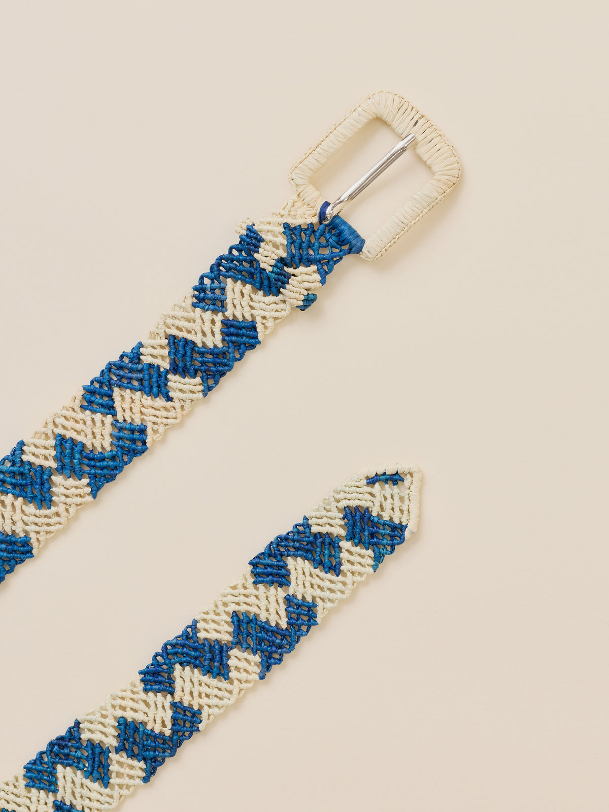 A close-up of a woven belt strap with a blue and cream chevron pattern, featuring a cream-colored metal buckle and a matching woven tip, set against a solid light beige background., image 2 of 2