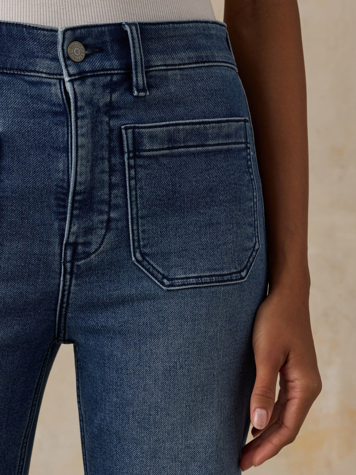 Close-up of a person wearing dark blue denim jeans, showing the back pocket, waistband, and hand against a warm beige background., image 5 of 6