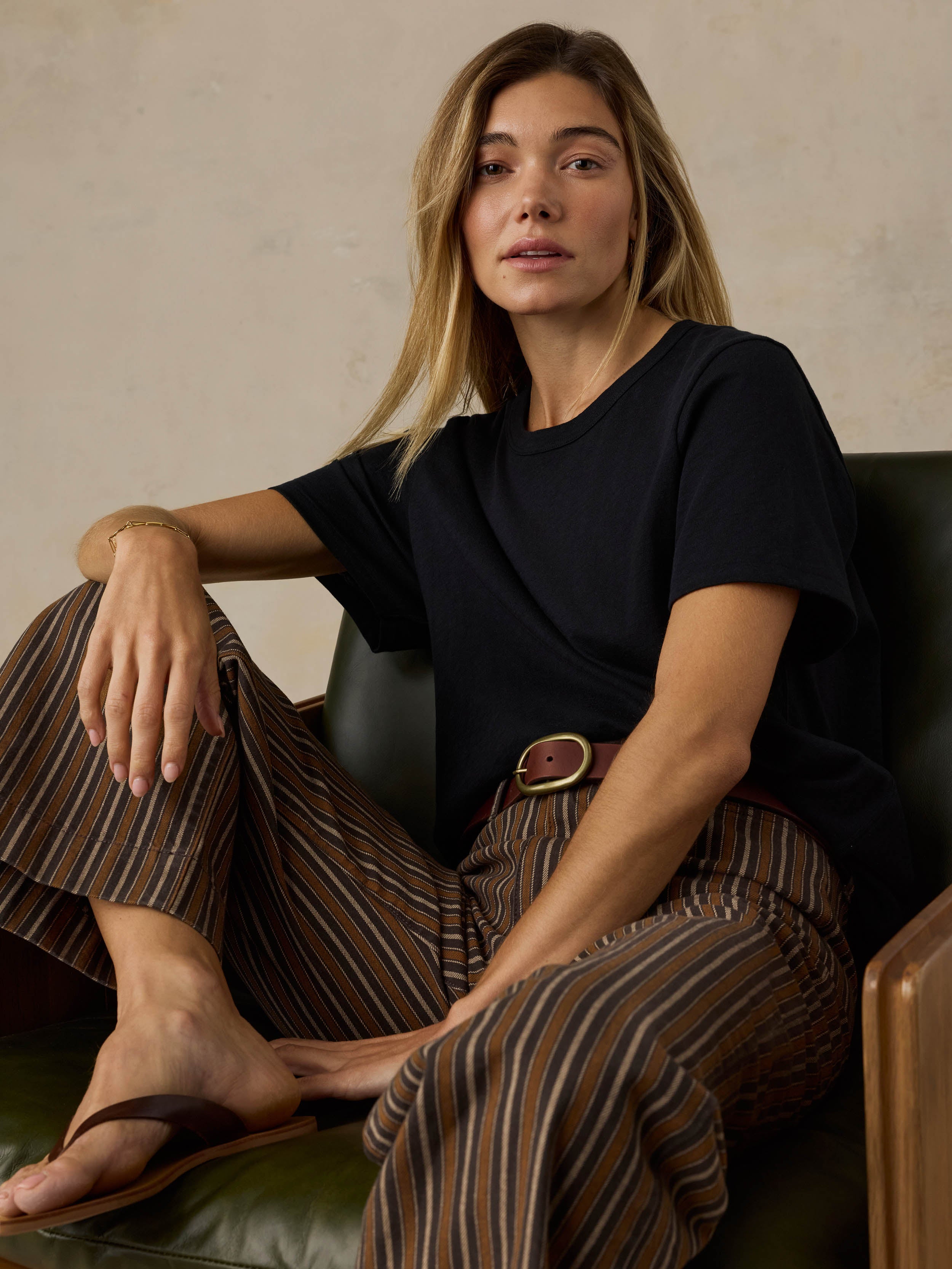 Woman sitting on a wooden chair wearing a black t-shirt and brown striped pants on a beige background