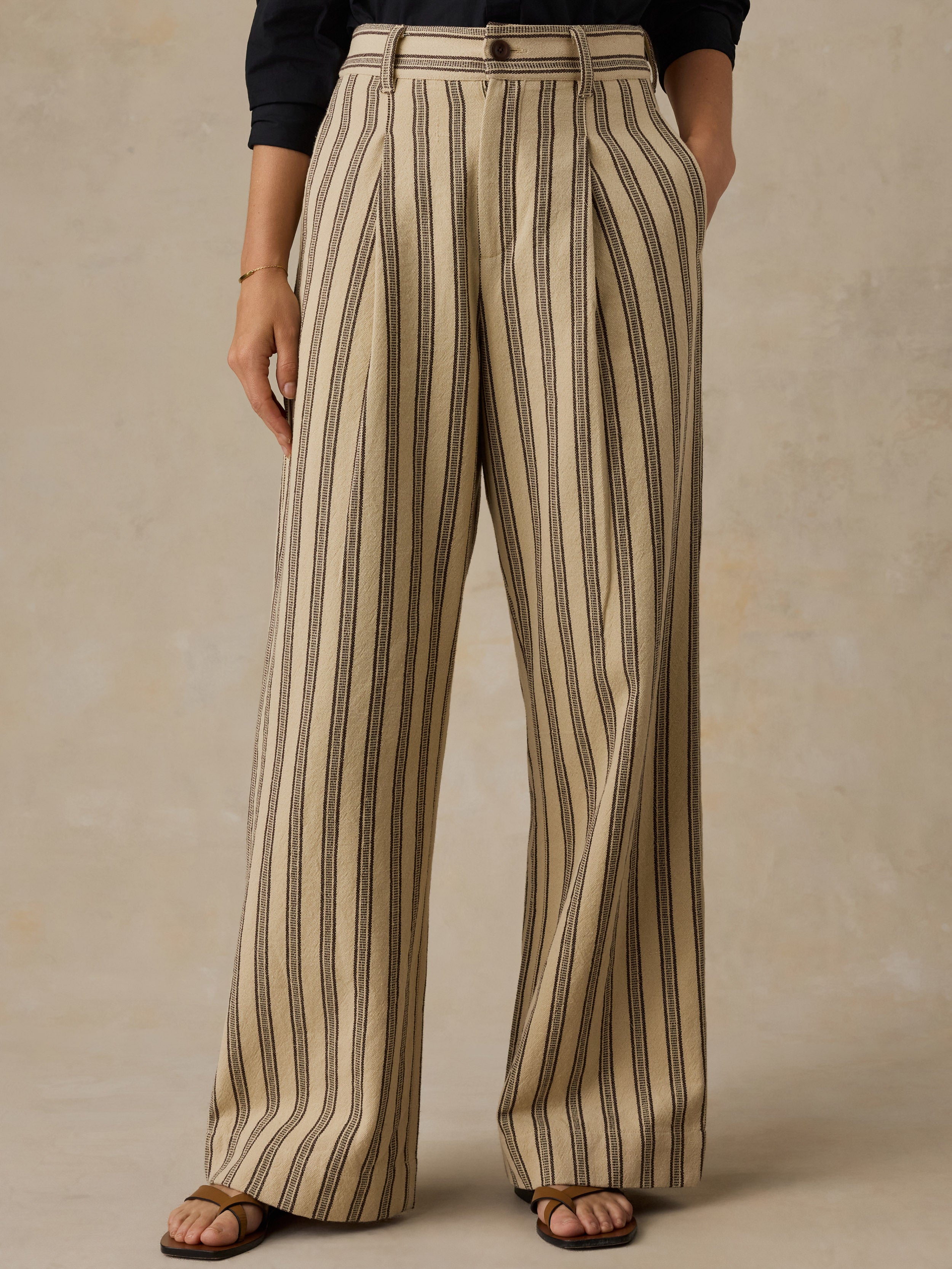 Woman wearing a black button down shirt and high waisted beige and black striped pants against a beige background.