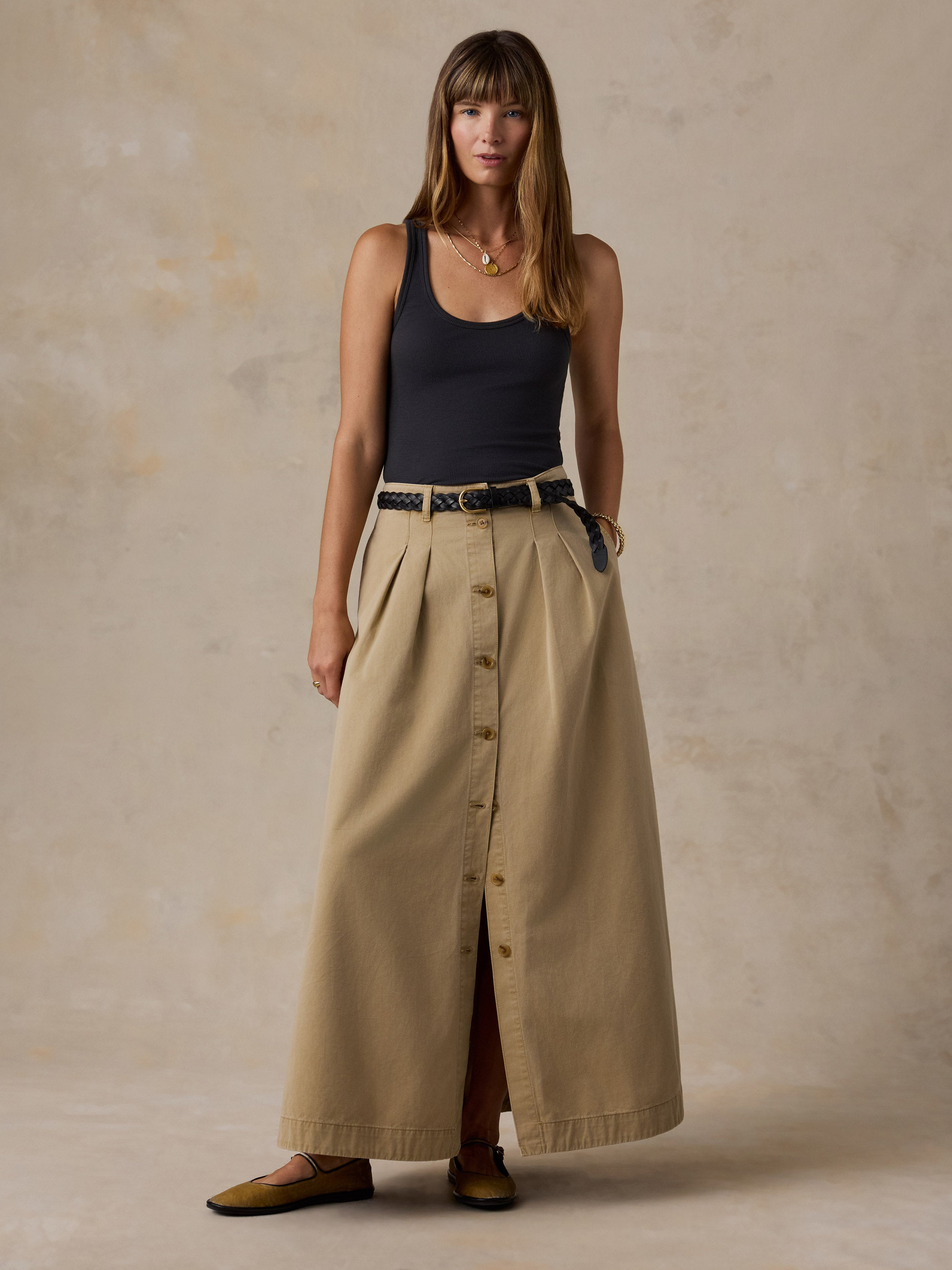 A woman stands against a textured beige studio backdrop wearing a black sleeveless top, a long beige button-front skirt with a black belt, and olive flats.