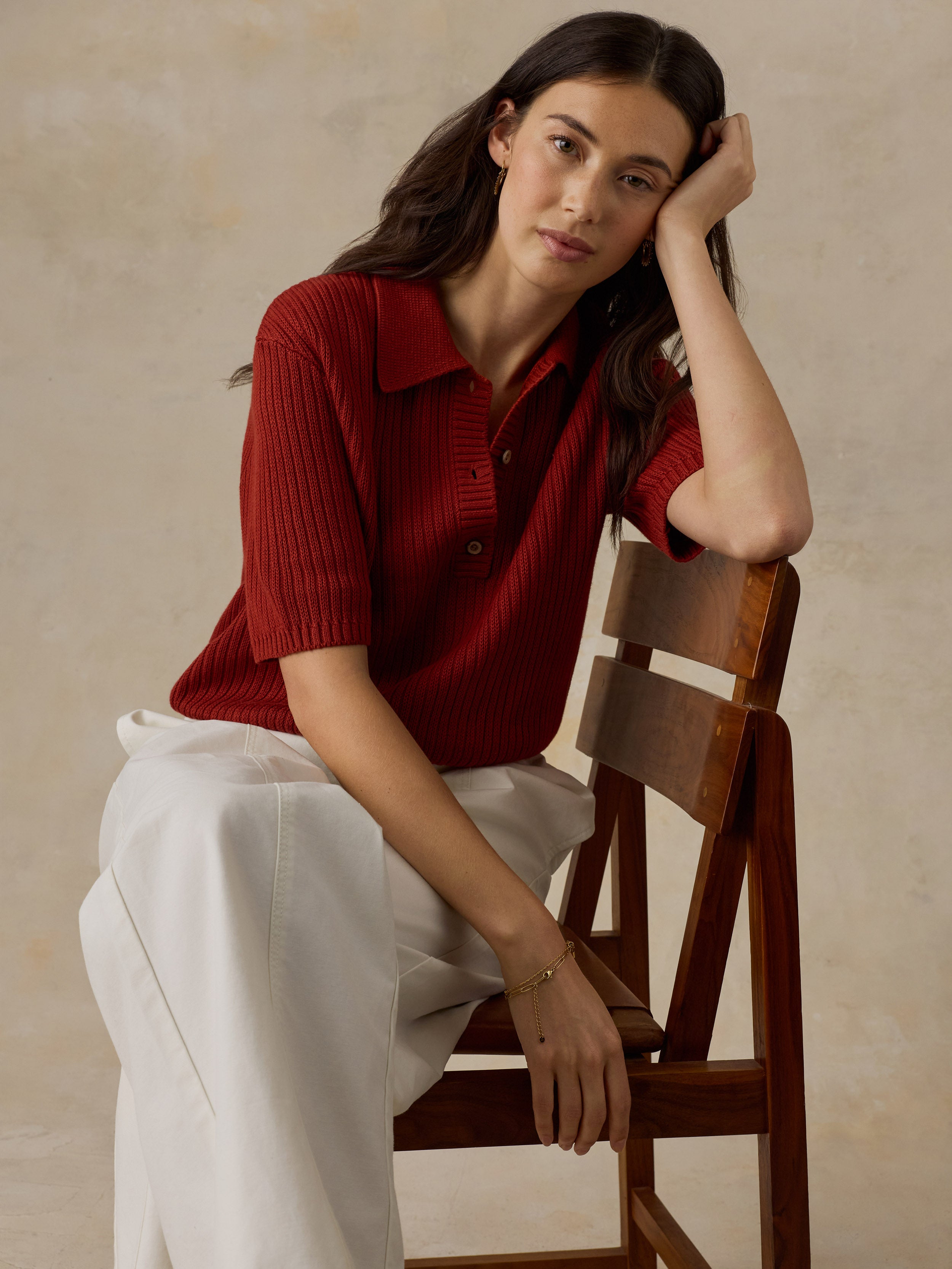 Woman sitting on a wooden chair wearing a red knit polo shirt and white drawstring pants against a beige background