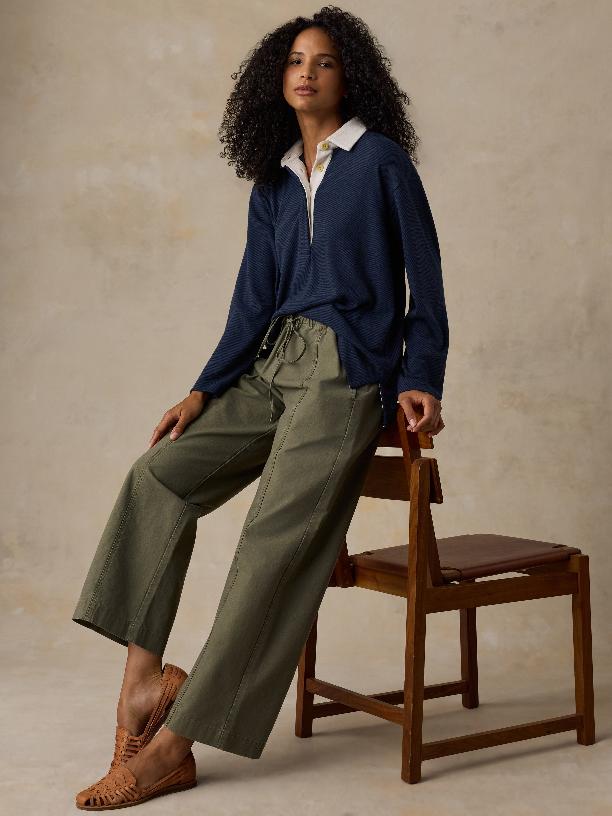 A woman with curly black hair sits on a wooden chair wearing a navy blue long-sleeve collared shirt, olive green wide-leg pants, and brown woven mule shoes against a textured beige studio backdrop.