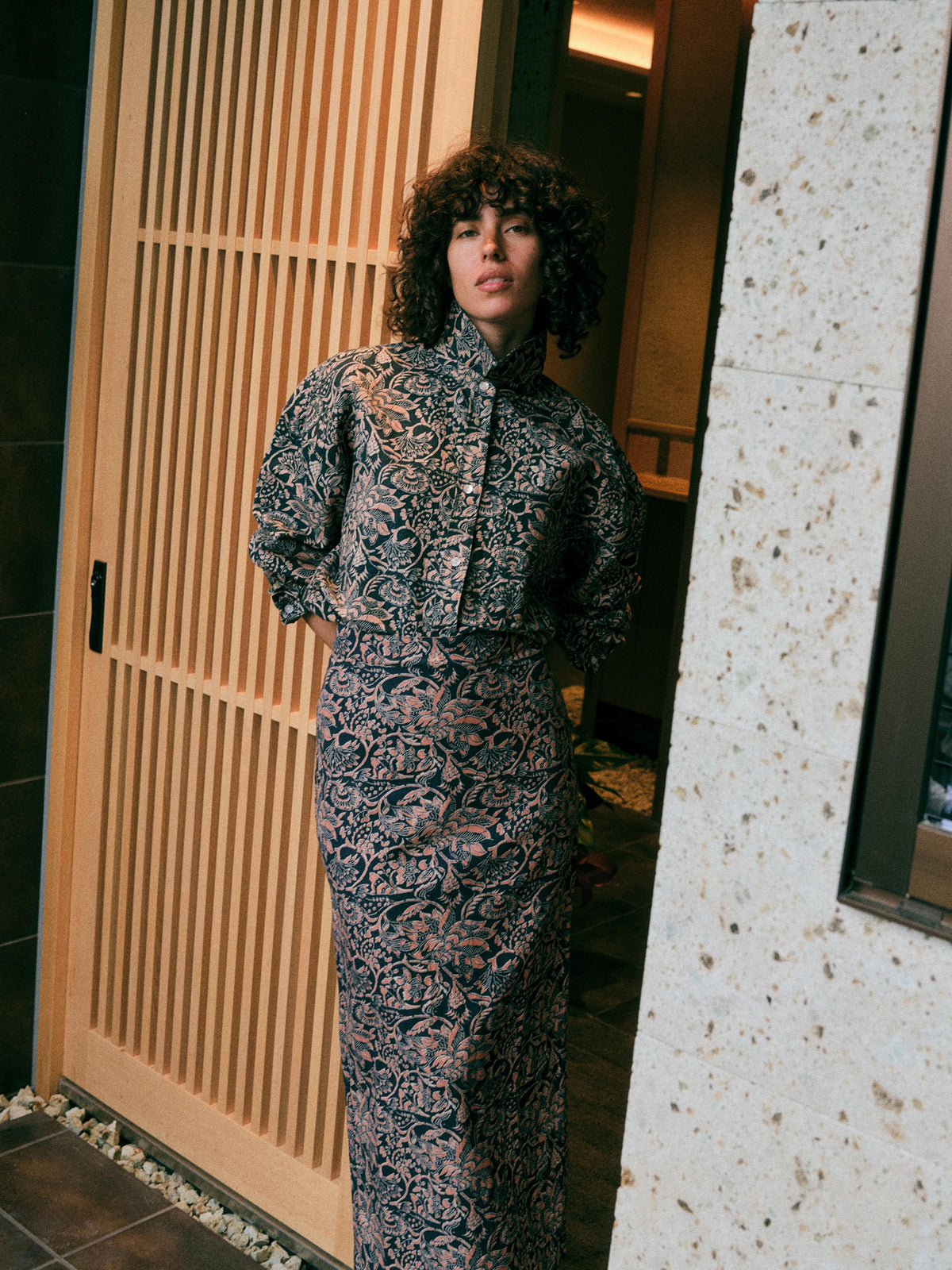 A woman with curly dark hair stands in a doorway wearing a long-sleeved, high-neck midi dress with a dense abstract floral pattern in black, navy, and dusty pink. The background features a light wood slatted door on the left and a textured stone pillar on the right., image 1 of 6