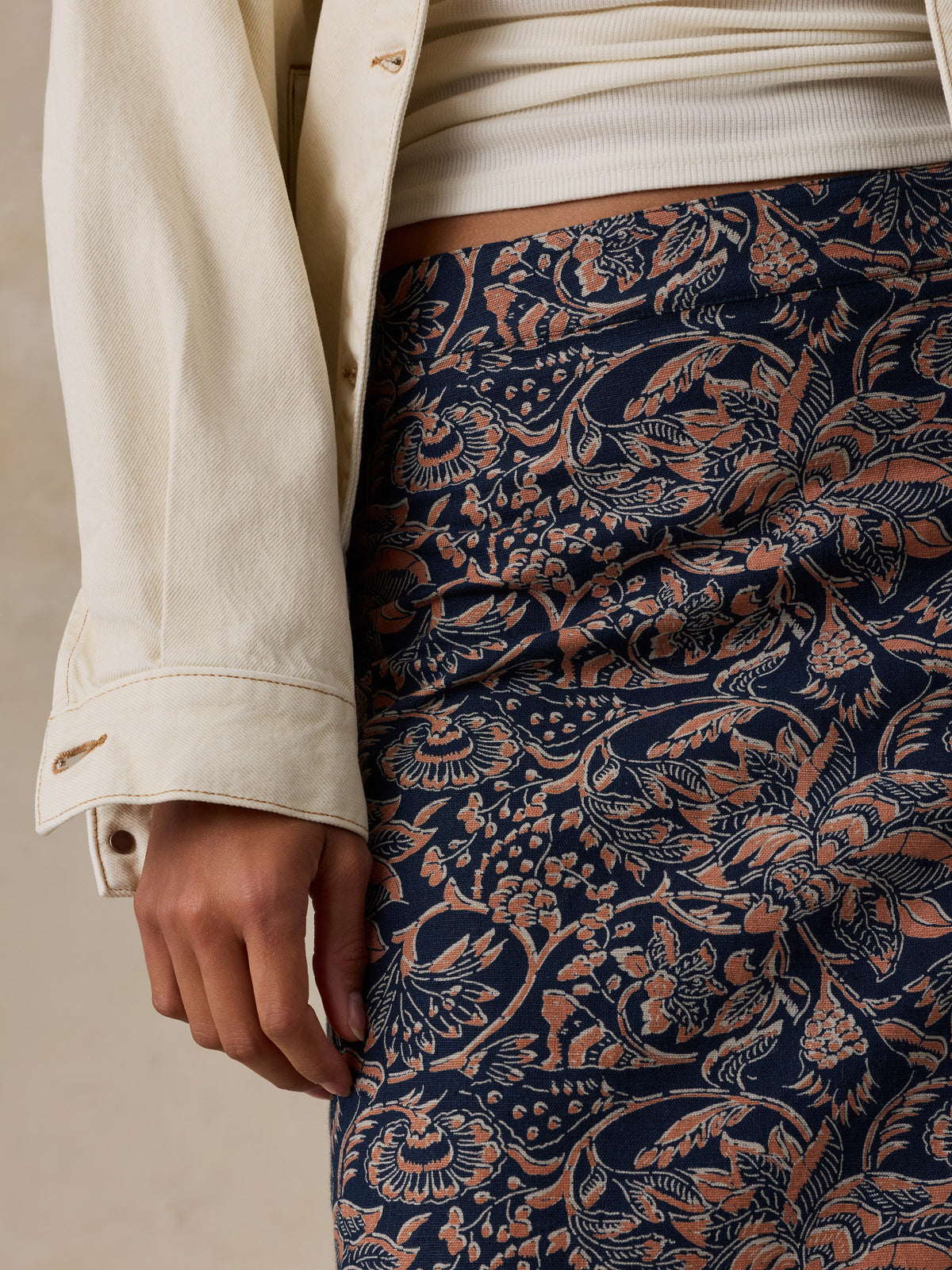 Close-up of a woman wearing a navy blue skirt with a detailed terracotta floral and botanical pattern, styled with a cream ribbed top and an off-white jacket., image 5 of 6