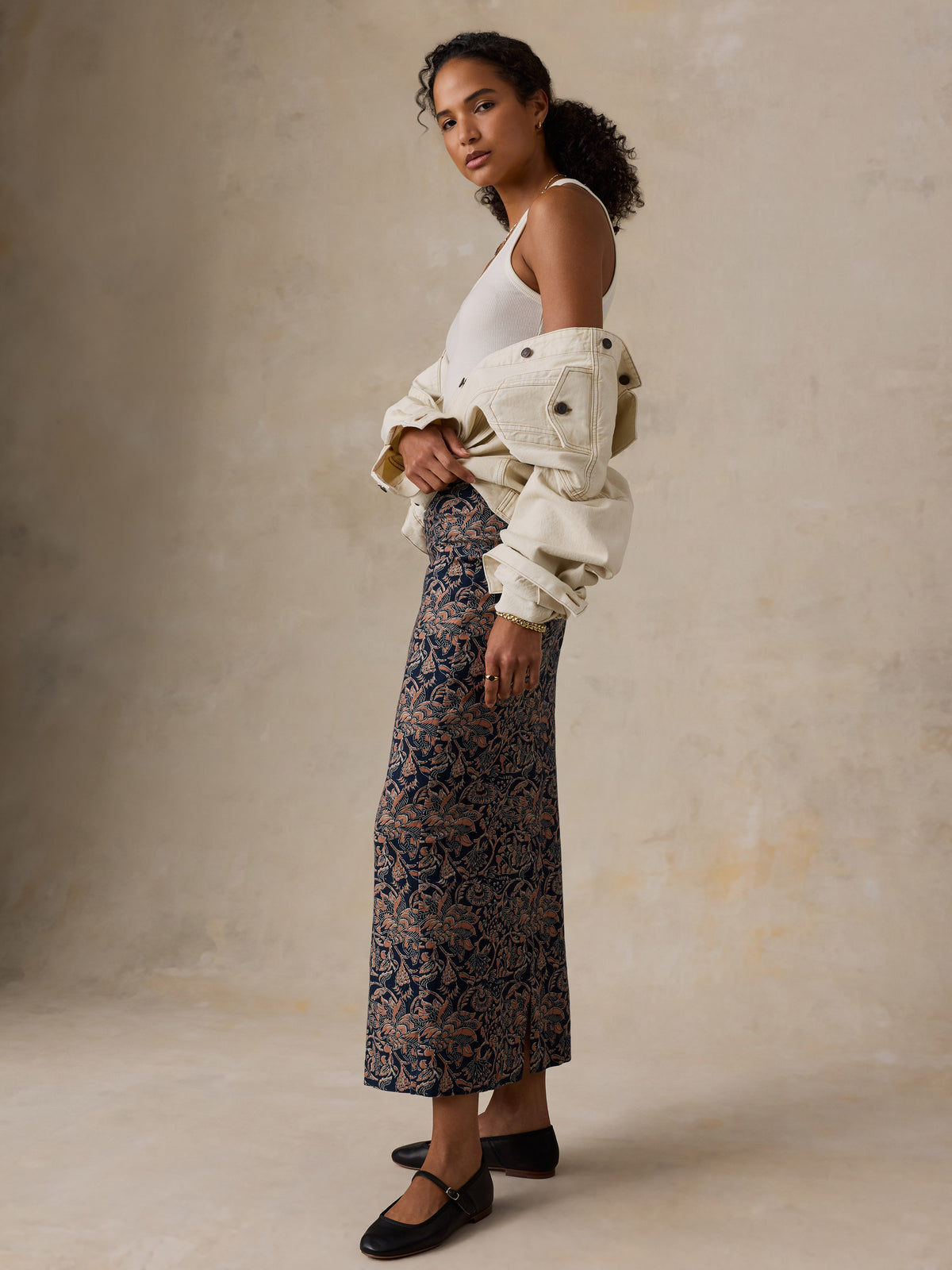 A woman models a long navy and beige patterned skirt paired with a white tank top, an off-white jacket draped off her shoulder, and black flats, set against a textured beige studio backdrop., image 3 of 6