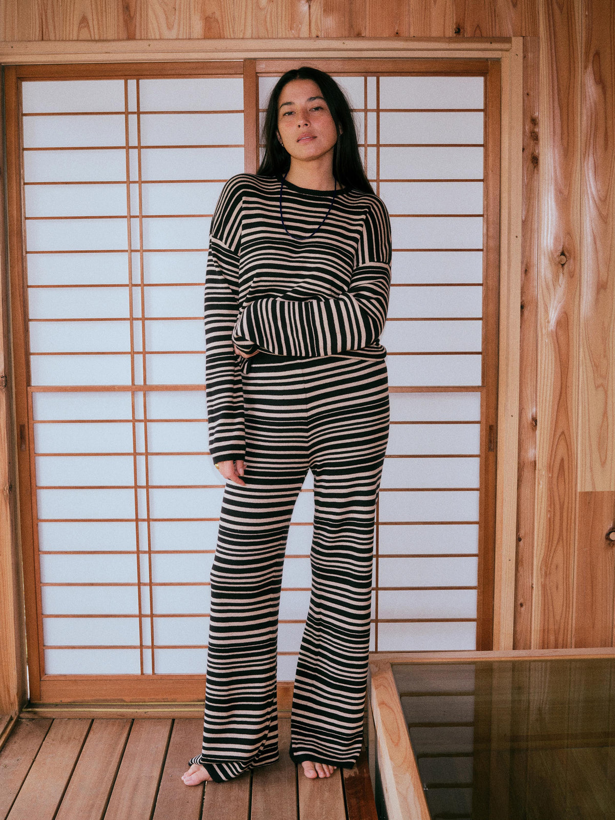 A woman stands barefoot in front of a wooden shoji screen, wearing a matching black and beige striped long-sleeve top and wide-leg pants set., image 1 of 6