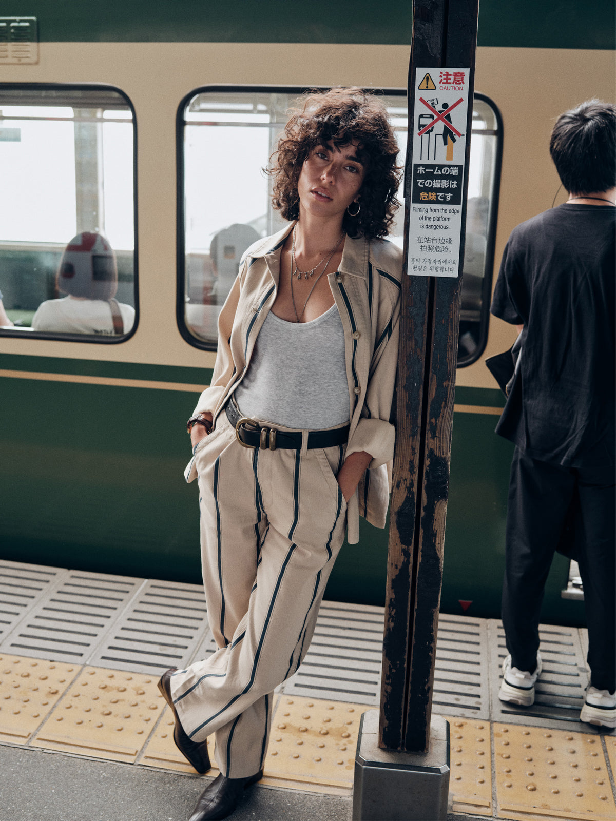 A woman stands on a subway platform wearing a beige striped suit with a white tank top and black belt, posing next to a train and a metal pole., image 8 of 8