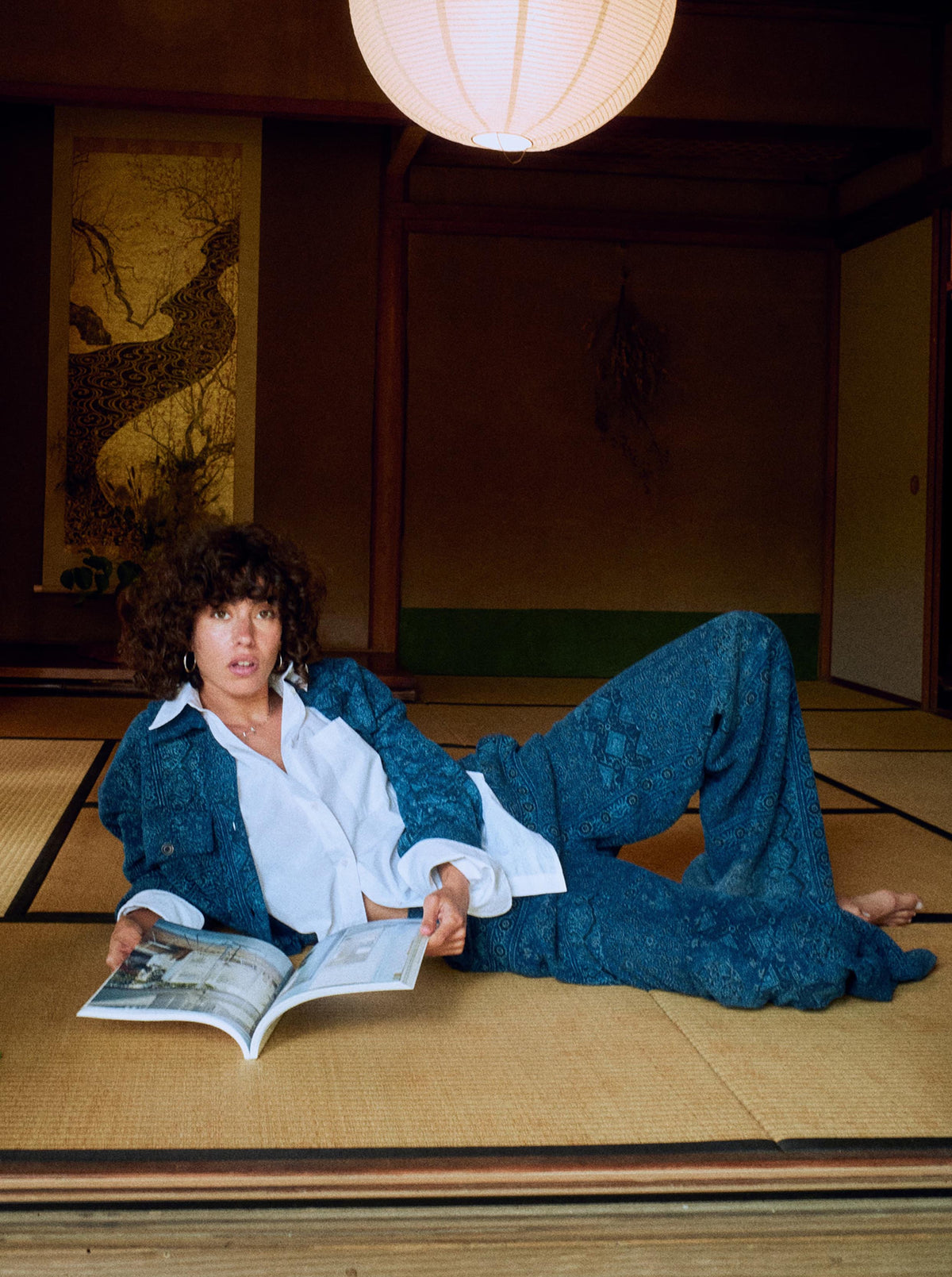 A woman reclining on a tatami floor in a traditional Japanese room, wearing a blue patterned jacket and matching wide-leg pants over a white shirt, while holding an open magazine., image 7 of 7