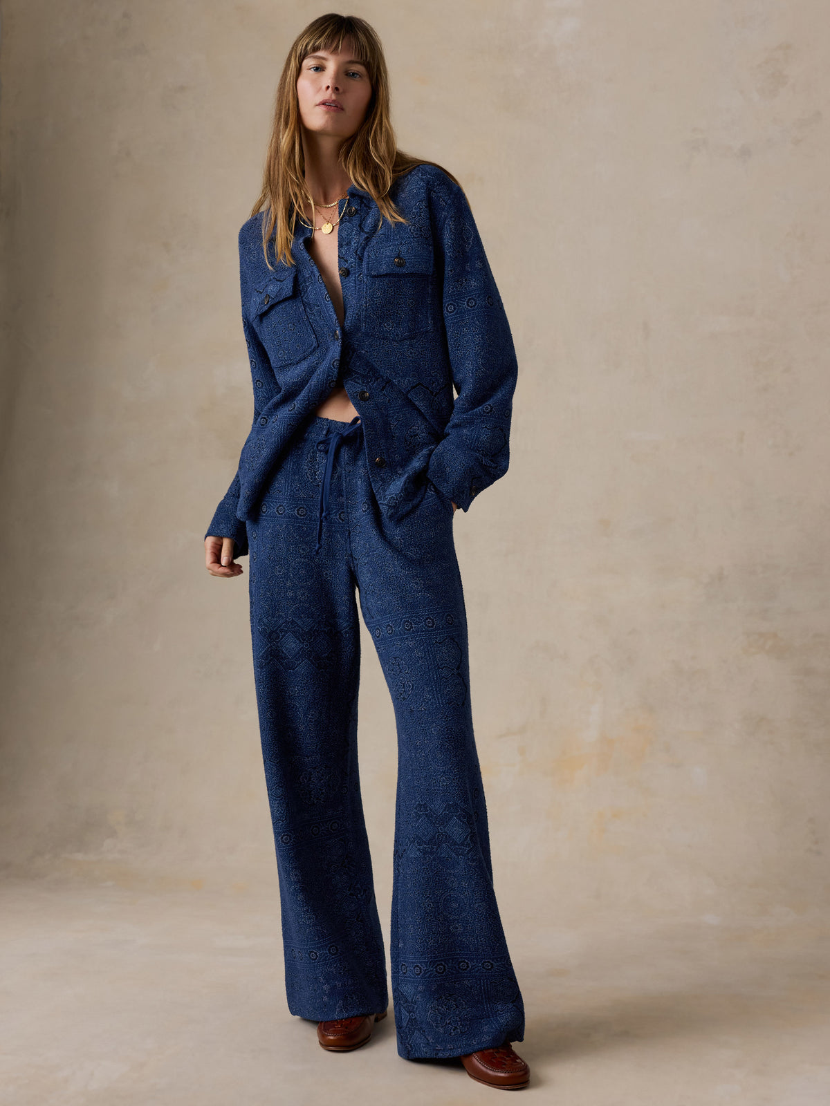 Woman wearing a printed pair of blue pants and matching top standing against a beige studio background, image 5 of 7
