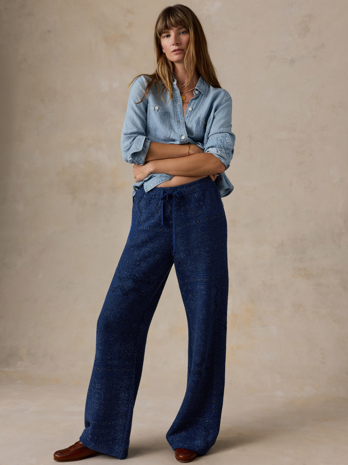 Woman wearing a printed pair of blue pants and a blue chambray shirt standing against a beige studio background, image 6 of 7
