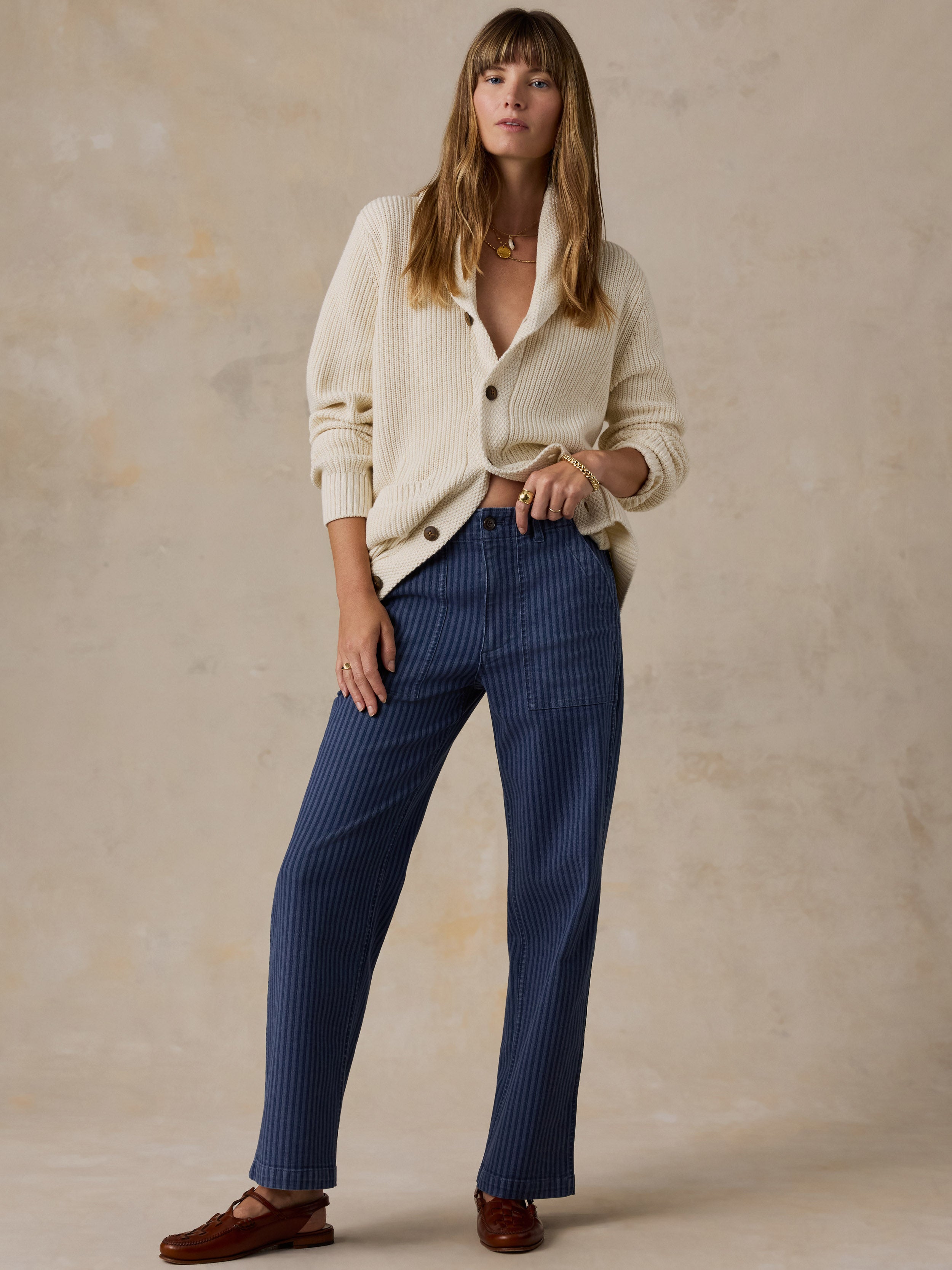 A woman stands against a textured beige studio backdrop wearing a cream ribbed cardigan, navy blue pinstriped trousers, and brown loafers.