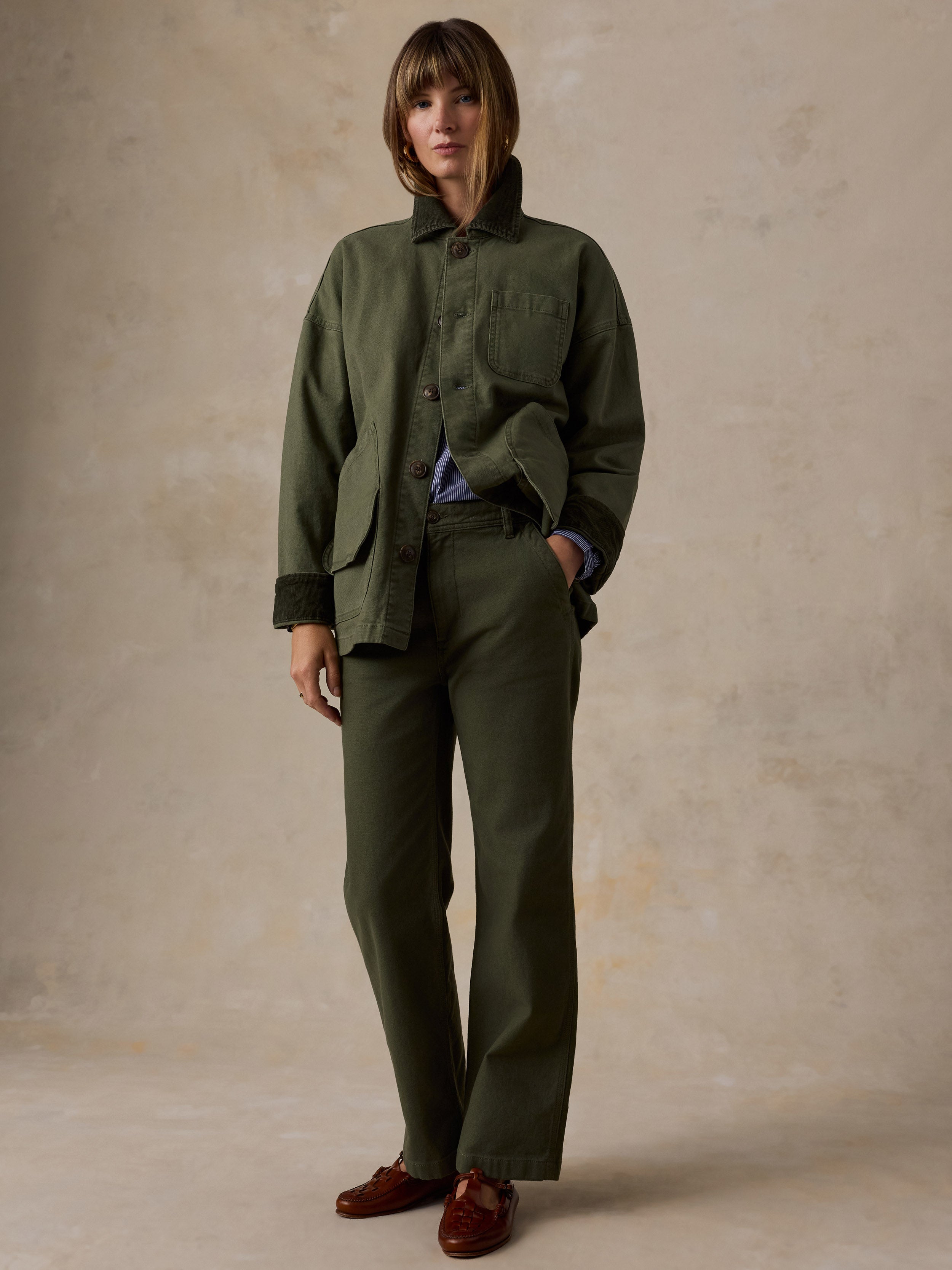 A model stands against a textured beige studio backdrop wearing an olive green military-style jacket and matching trousers, layered over a blue shirt and paired with brown leather loafers.