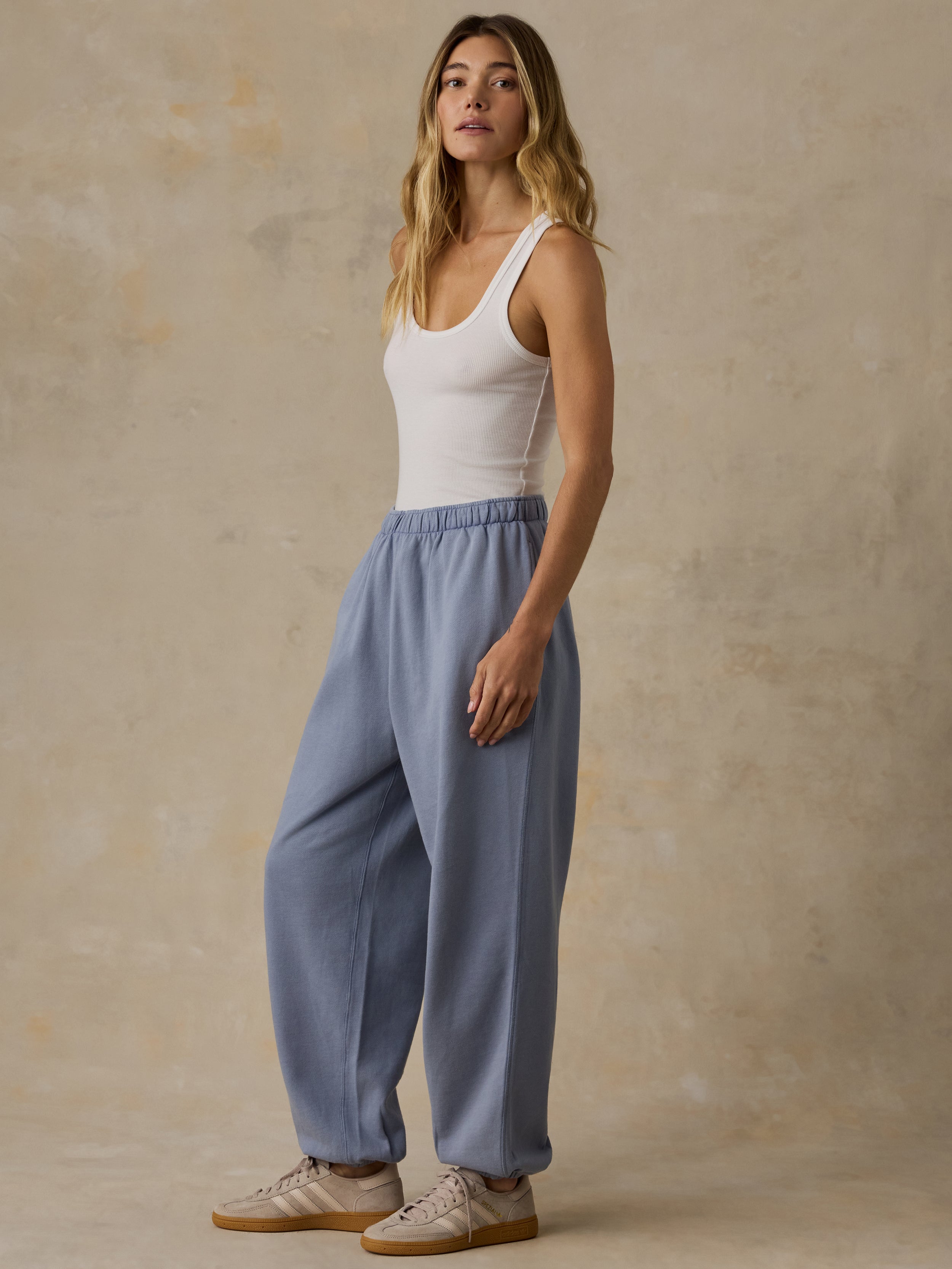 Woman wearing a white tank top and blue sweatpants against a beige background