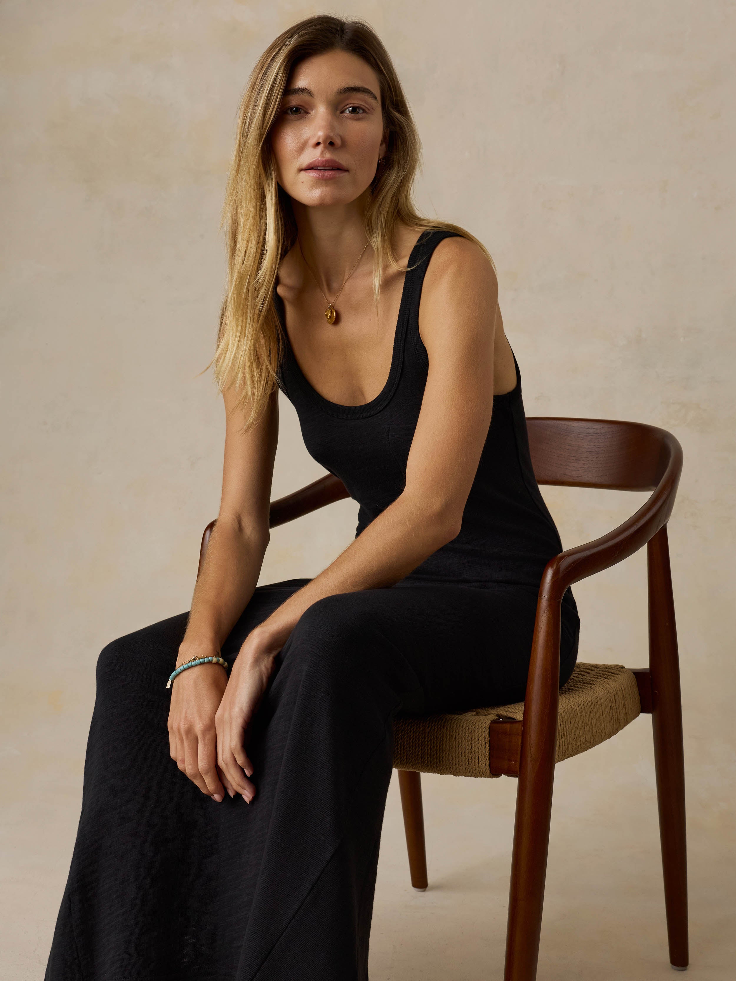 Woman sitting on a wooden chair wearing a black sleeveless maxi dress against a beige background