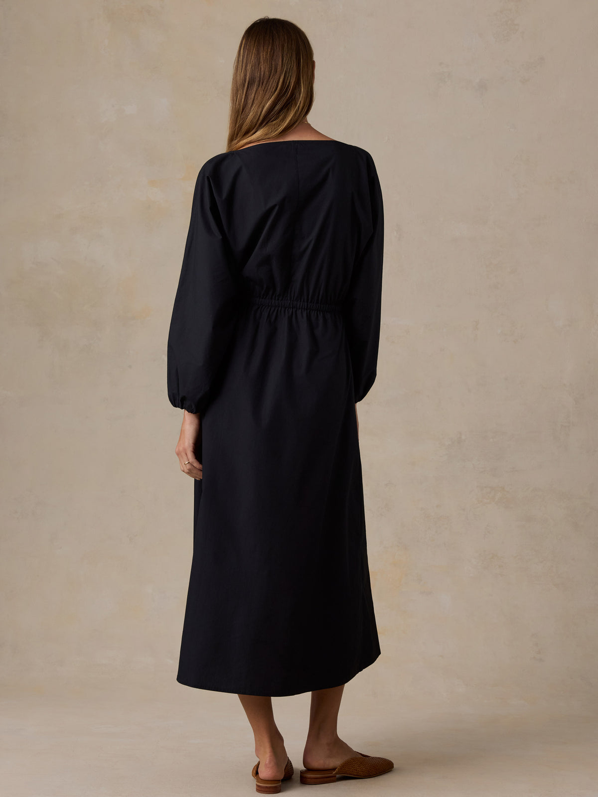 A woman viewed from the back wearing a long black midi dress with balloon sleeves and a gathered waist, standing against a warm beige textured studio backdrop., image 3 of 4