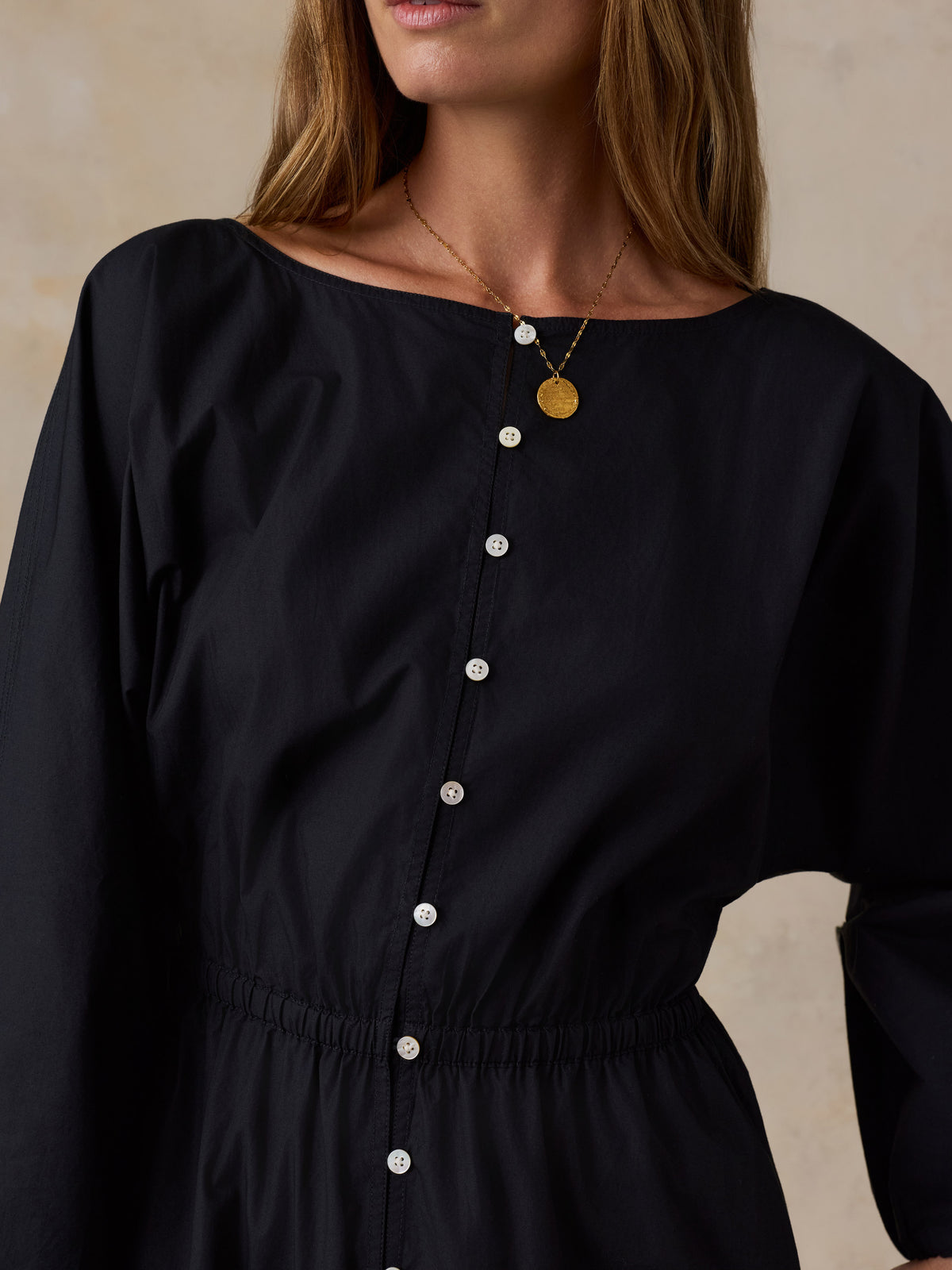 A close-up of a woman wearing a black long-sleeve dress with a boat neckline, front button closure, and elasticized waist, styled with a gold pendant necklace against a neutral beige background., image 2 of 4