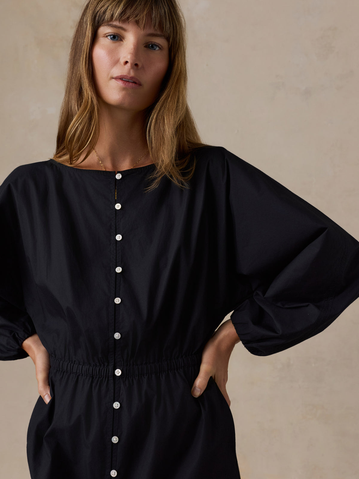 A woman wearing a black long-sleeve button-front dress or romper with a gathered elastic waist, posed against a warm beige textured wall., image 4 of 4