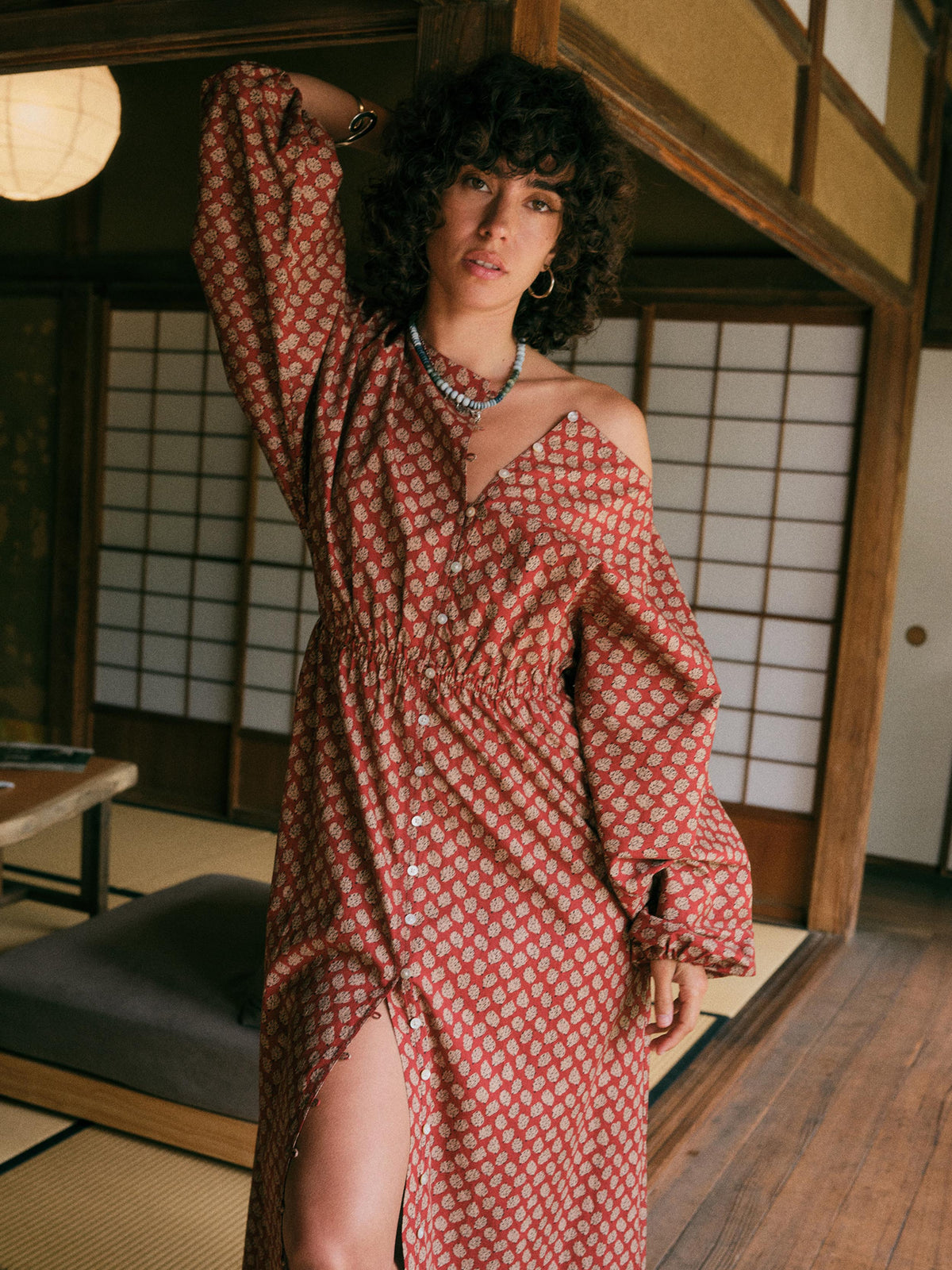 A woman with curly dark hair models a rust-red off-the-shoulder maxi dress with a white floral pattern, set in a traditional Japanese room with shoji screens., image 1 of 6