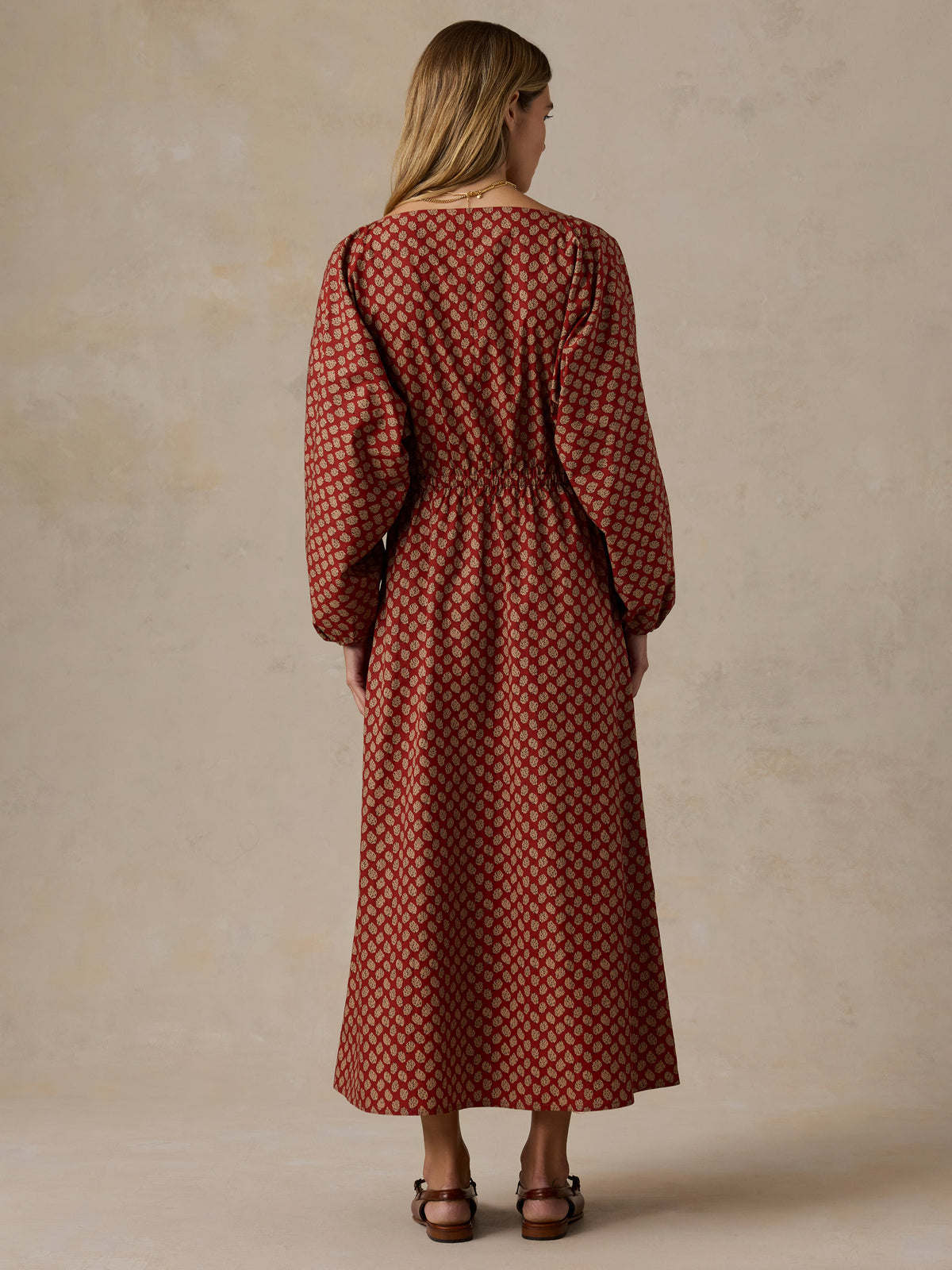 A woman viewed from the back wearing a long-sleeved midi dress with a rust-red and brown geometric pattern, standing against a textured beige studio backdrop., image 5 of 6
