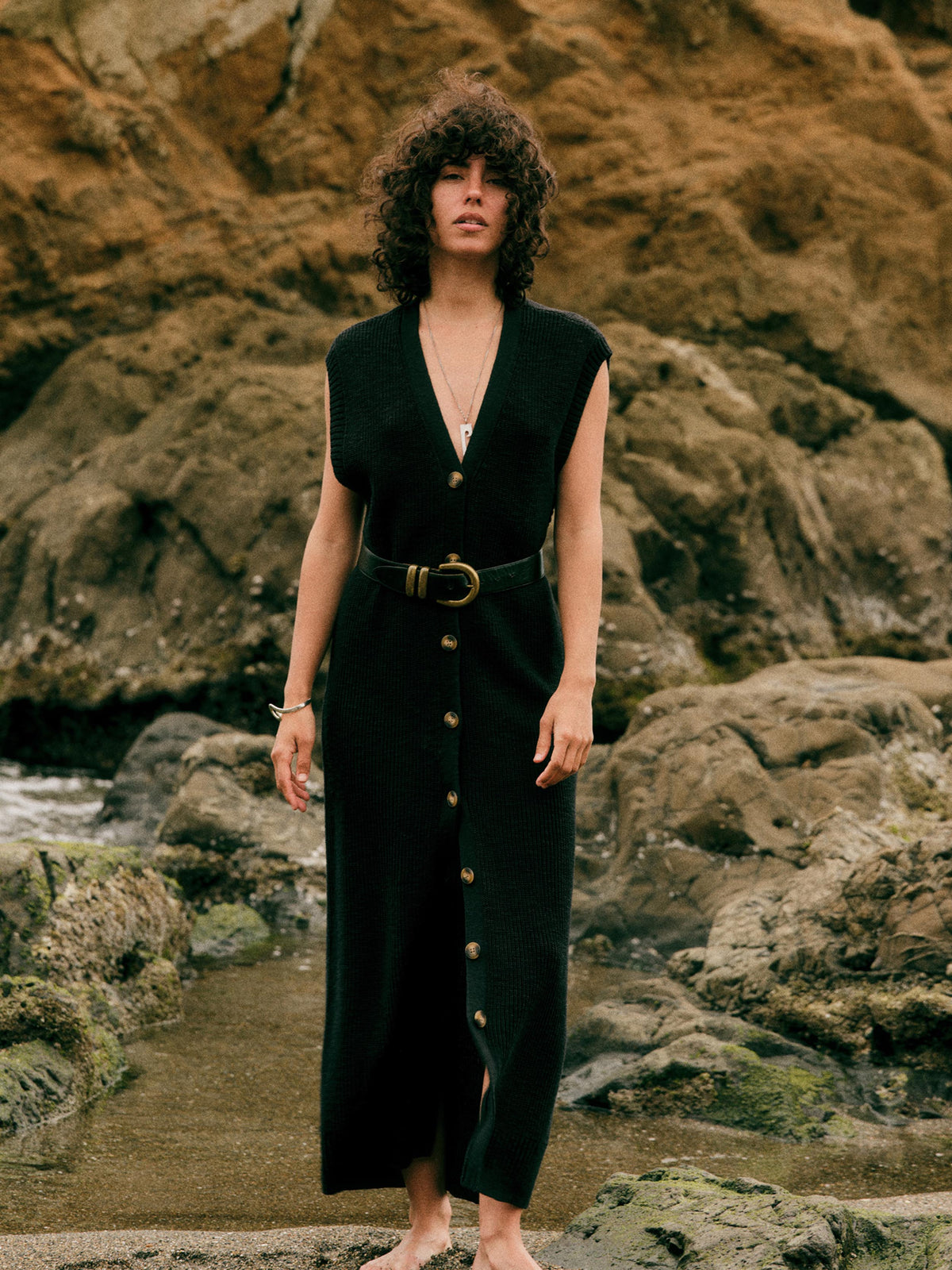A woman with curly dark hair stands barefoot on rocky coastal ground wearing a sleeveless black maxi dress with a V-neck, front button closure, and a matching belt with a large round buckle., image 1 of 5